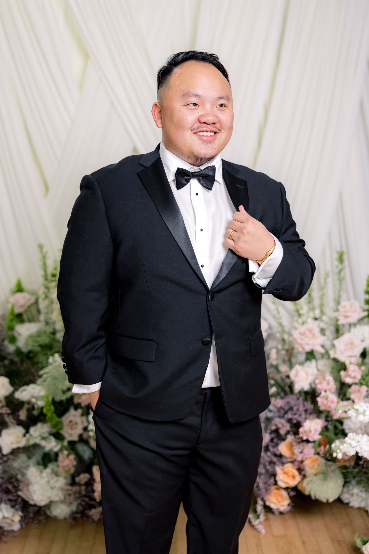 A formal portrait with classic black tuxedo and bow tie against a floral backdrop.