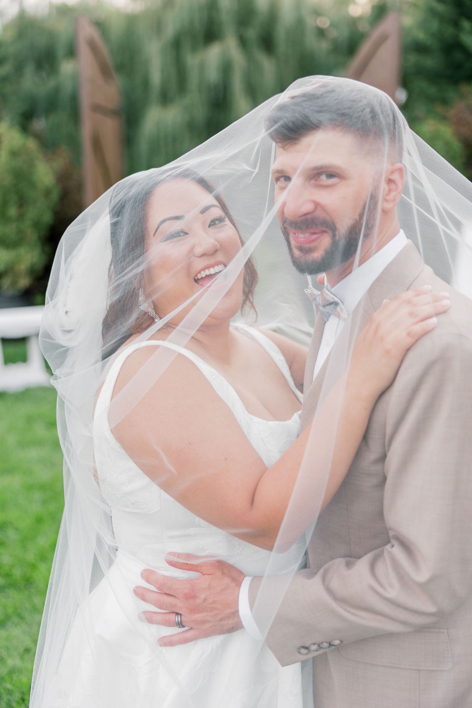 Joyful wedding couple sharing intimate moment under flowing veil outdoors.