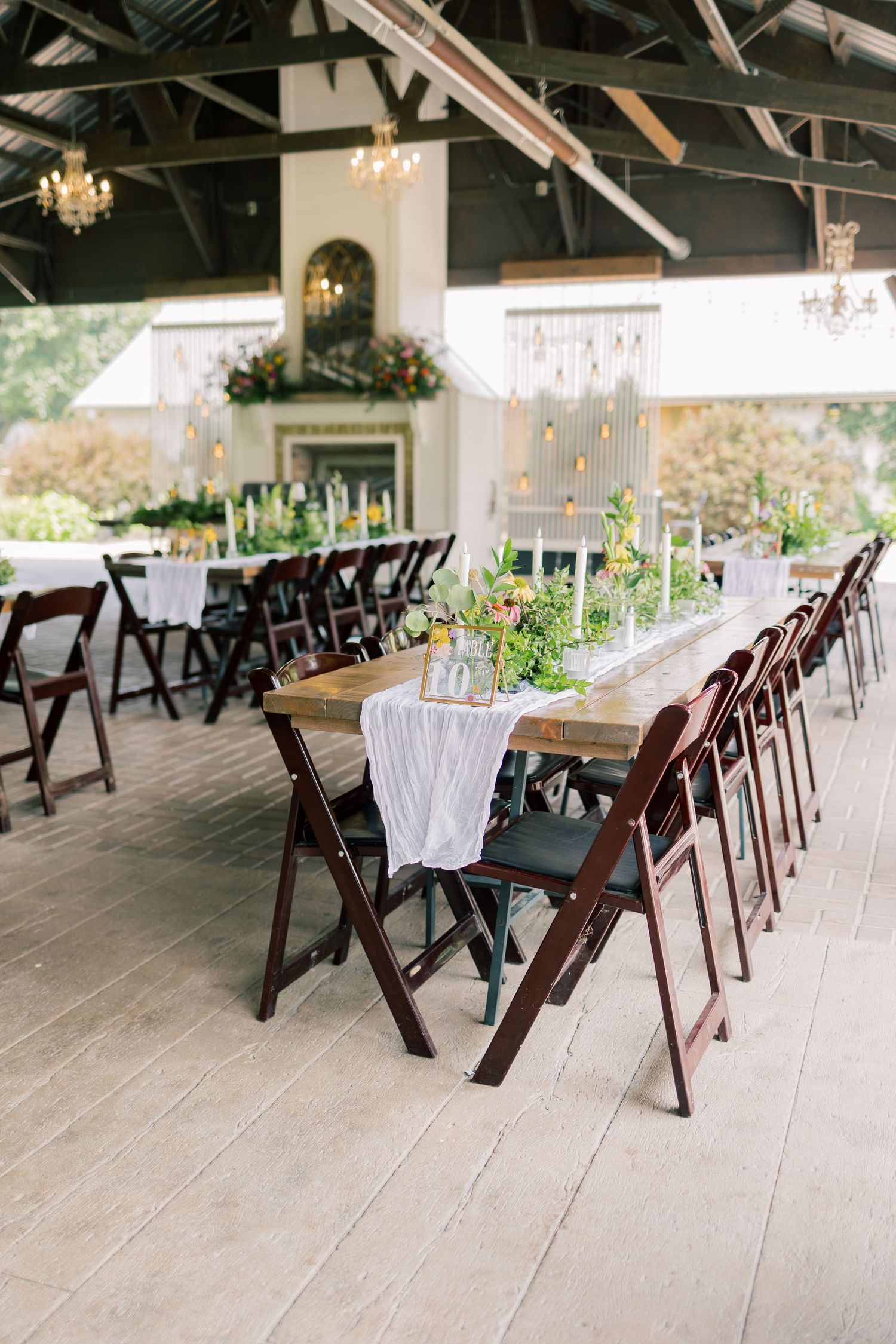 Wedding reception setup with long wooden tables, dark chairs, and floral centerpieces under a vaulted ceiling.