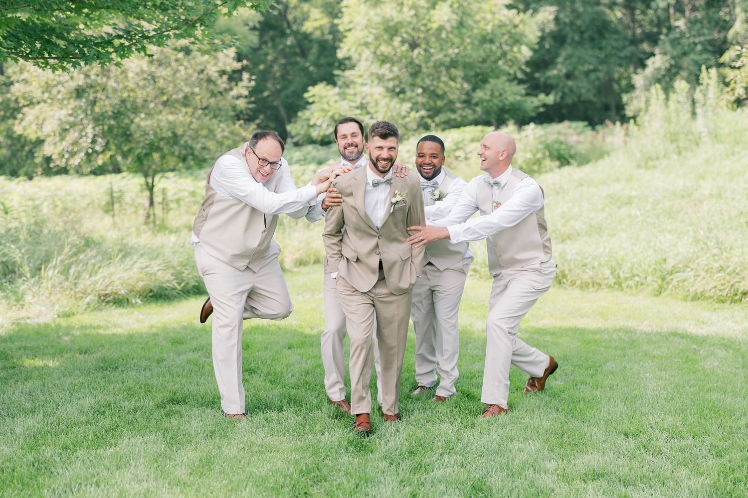 Groomsmen in tan suits playfully pose on a sunny grassy lawn during wedding celebrations.