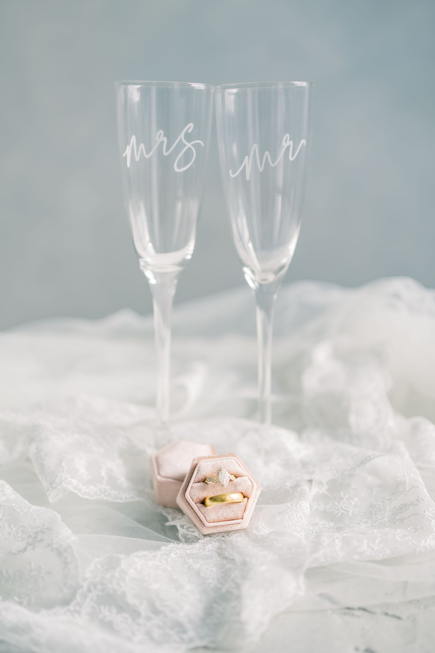 Two elegant champagne flutes with 'mrs' and 'mr' text sit beside a pink ring box on white lace fabric.