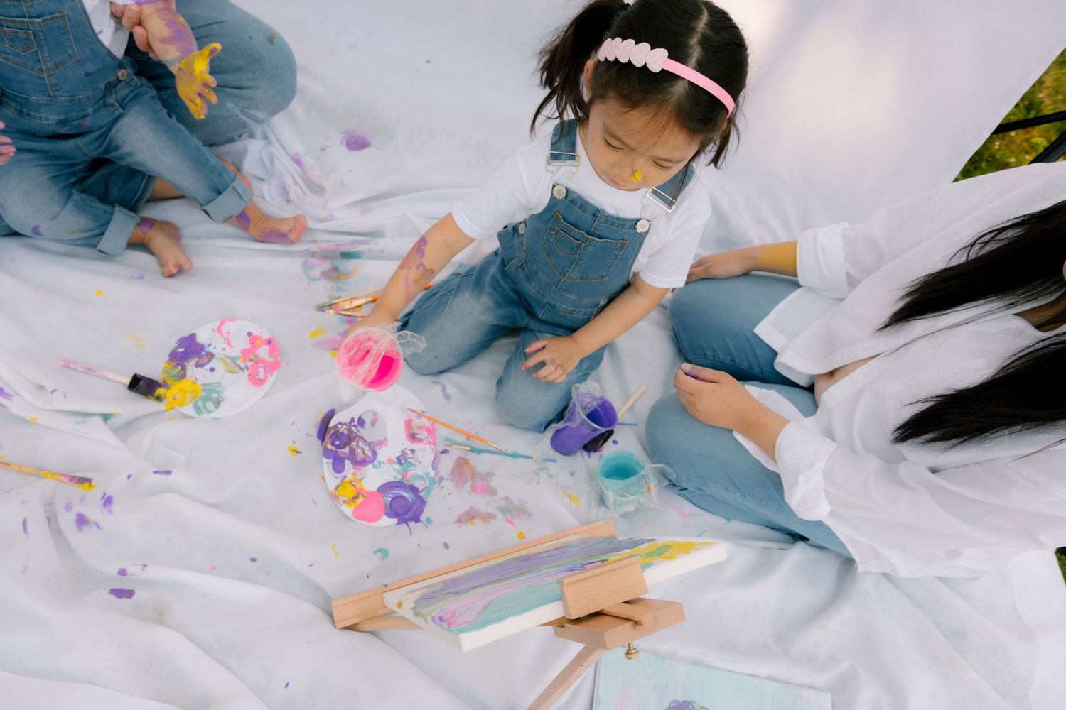 Young girl in denim overalls and mask painting colorfully on white canvas outdoors with art supplies around her.
