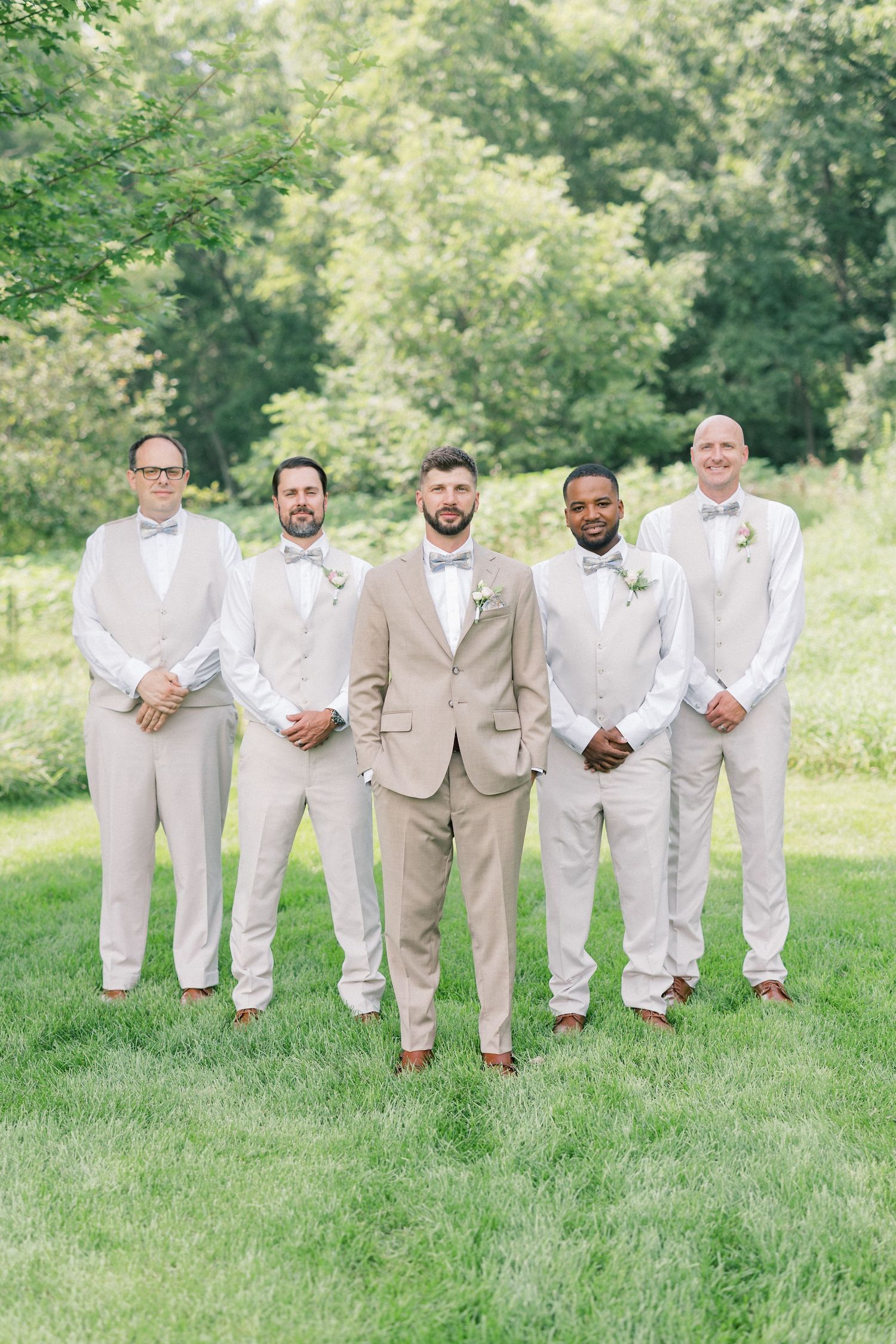 A group of groomsmen in light beige and white suits pose playfully on grass during an outdoor wedding photoshoot.