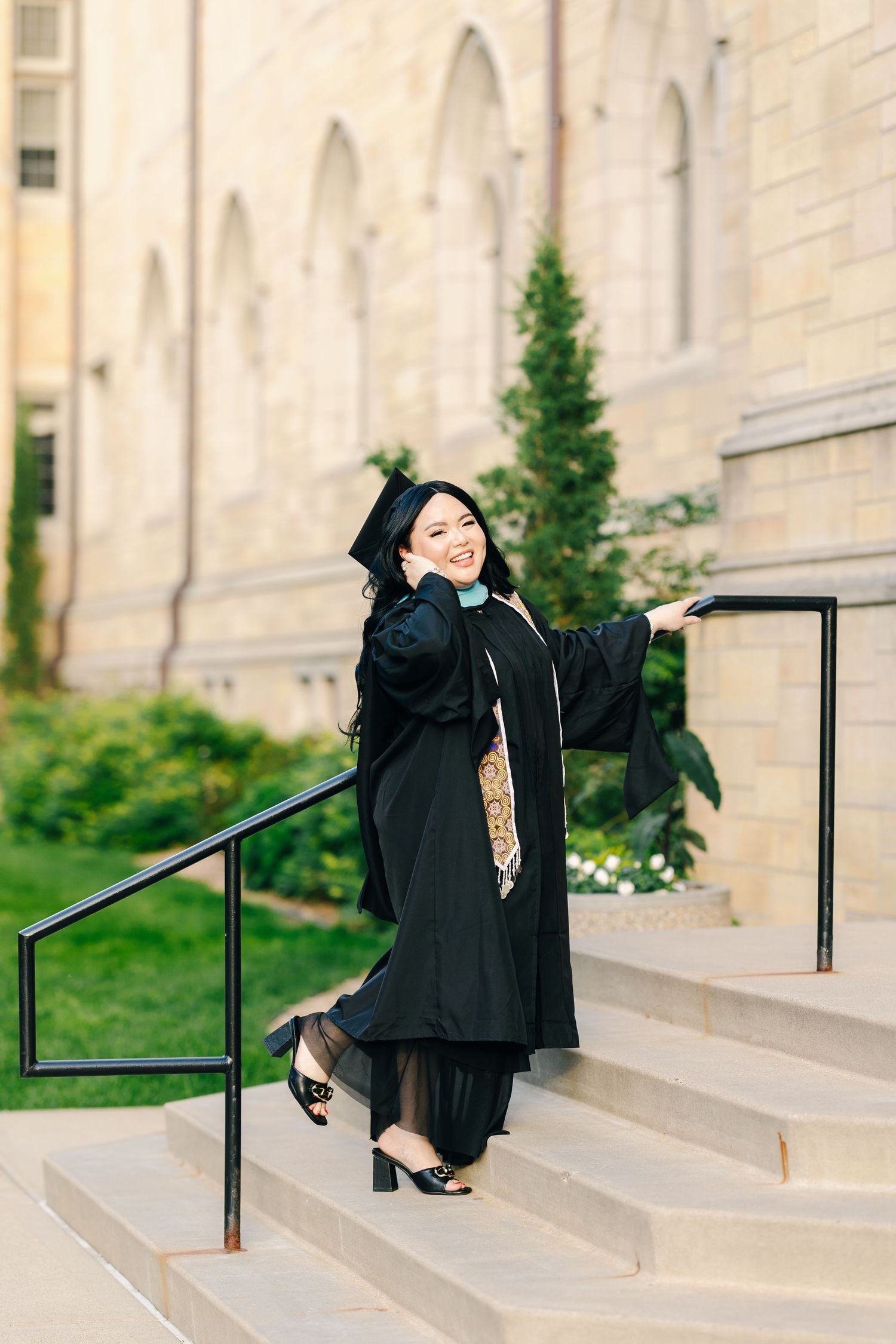 Graduate in black cap and gown posing joyfully on stone steps of a university building with gothic arches.