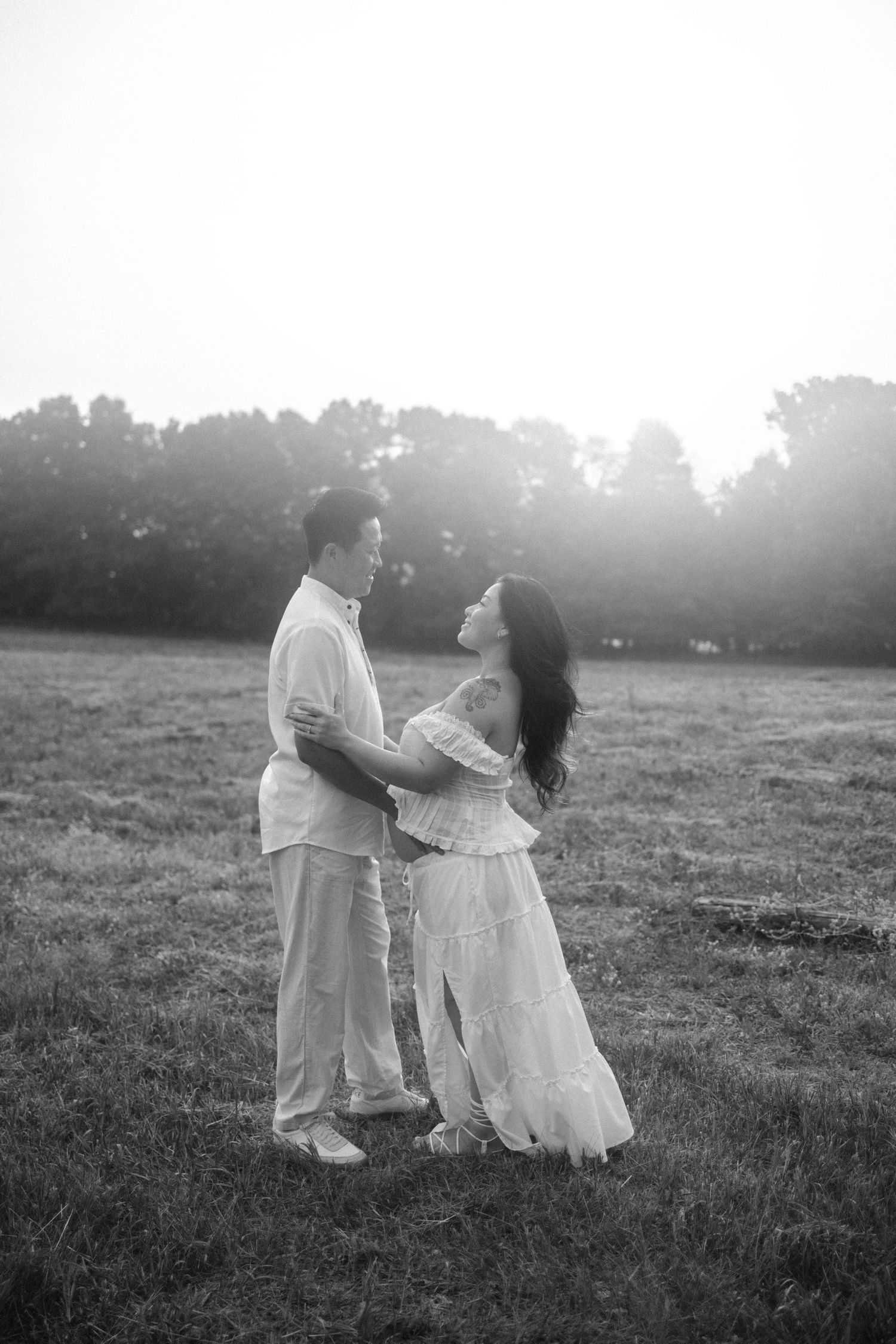 Black and white photo of a couple embracing in a sunlit field with trees in the background.