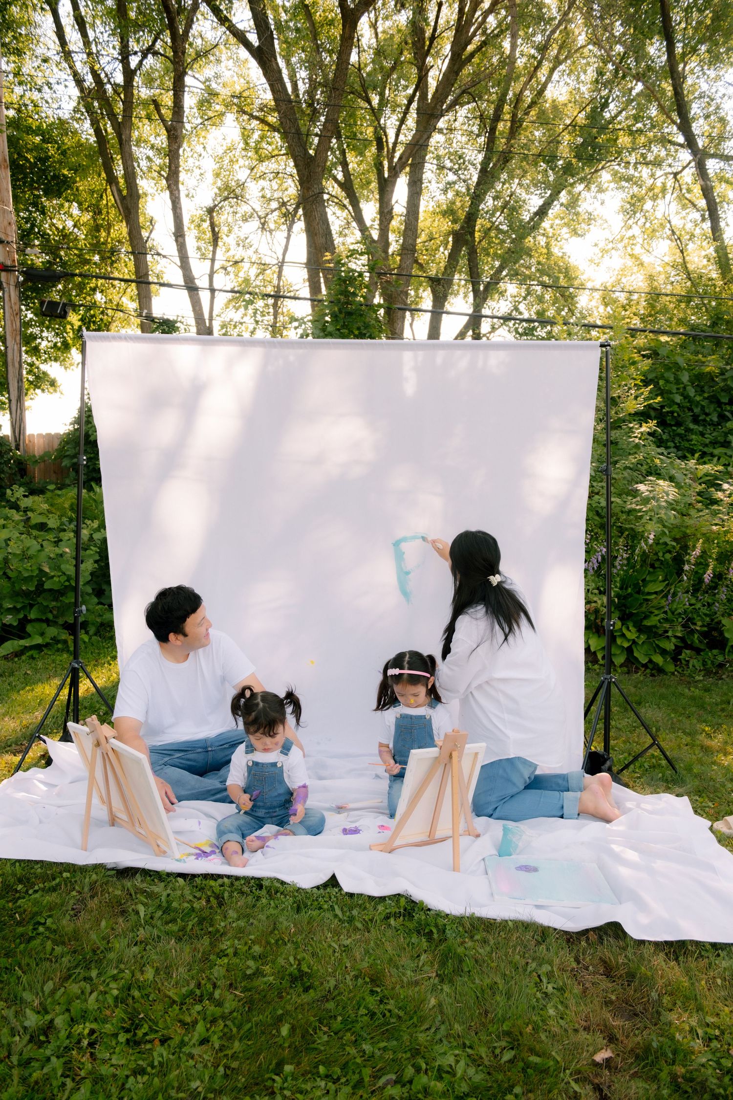 Asian family with two young daughters painting outdoors on white blanket with backdrop in garden.