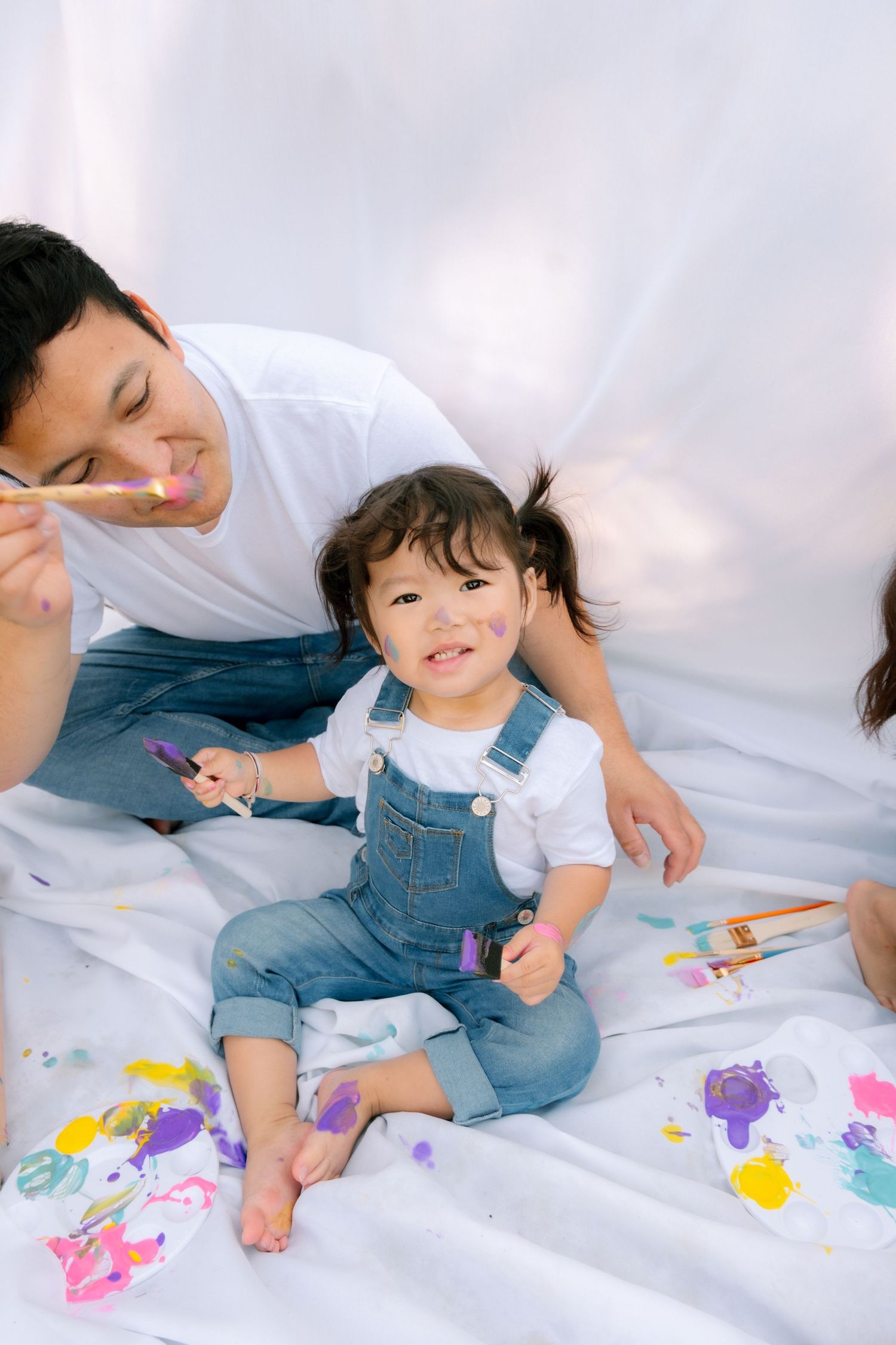 Father and toddler daughter enjoy painting together on a white backdrop outdoors.