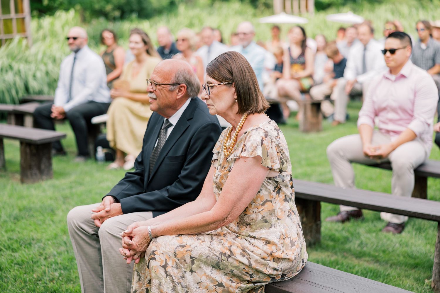 Wedding guests seated on wooden benches during outdoor ceremony.
