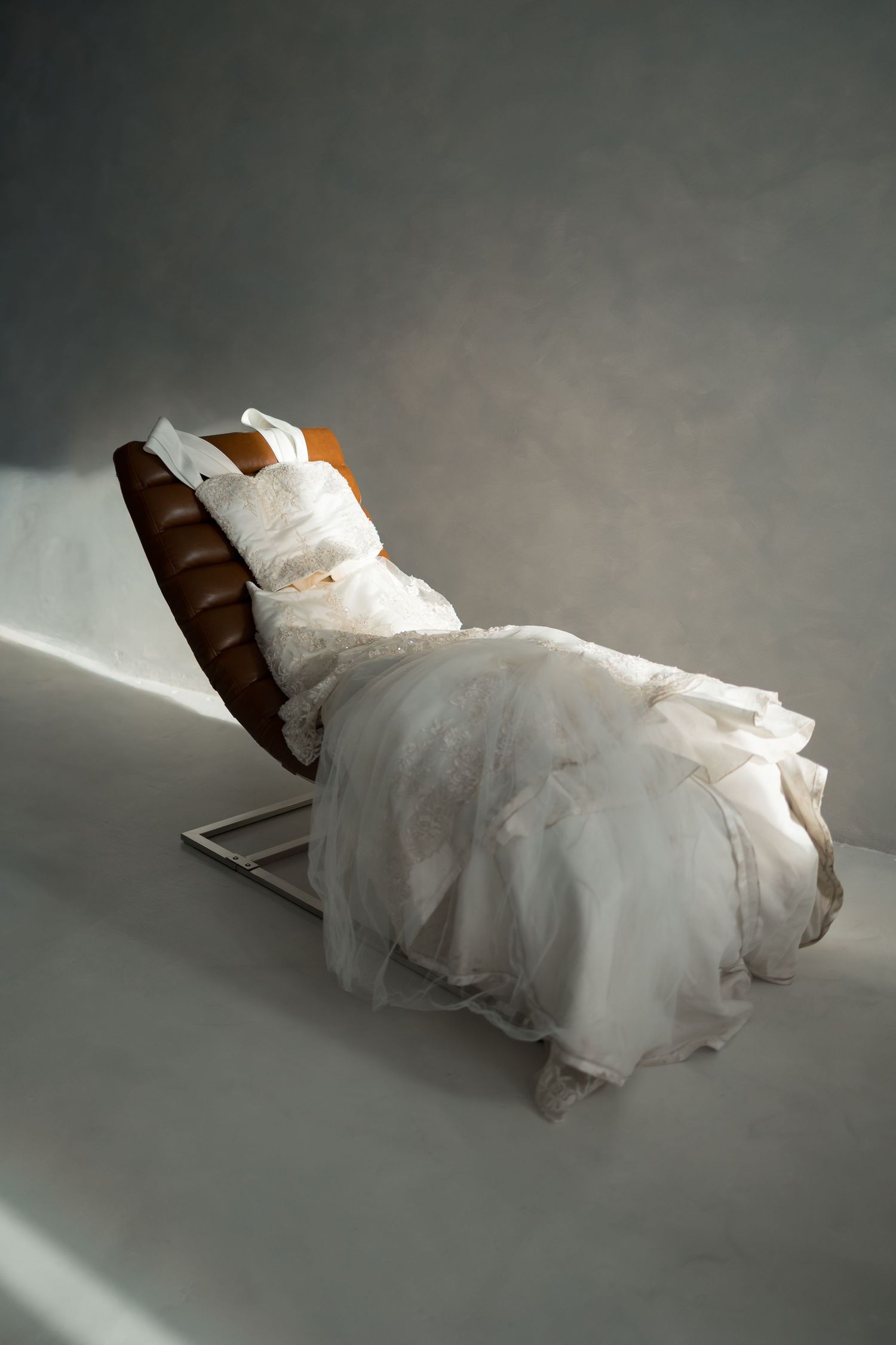 White wedding gown elegantly arranged on a brown leather reclining chair in a studio setting.