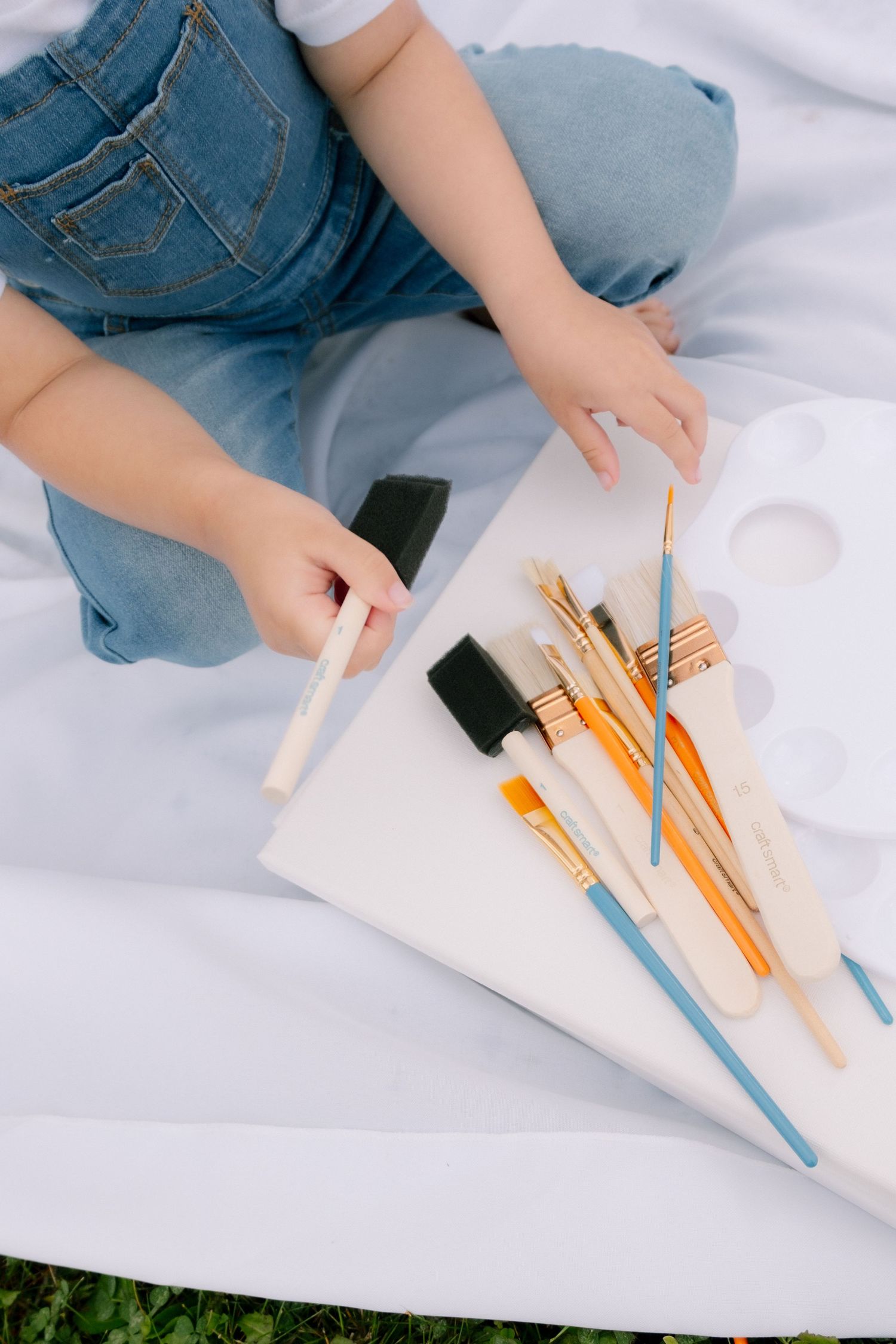 Child in denim overalls sits on white blanket outdoors, holding a sponge brush near art supplies and canvas.