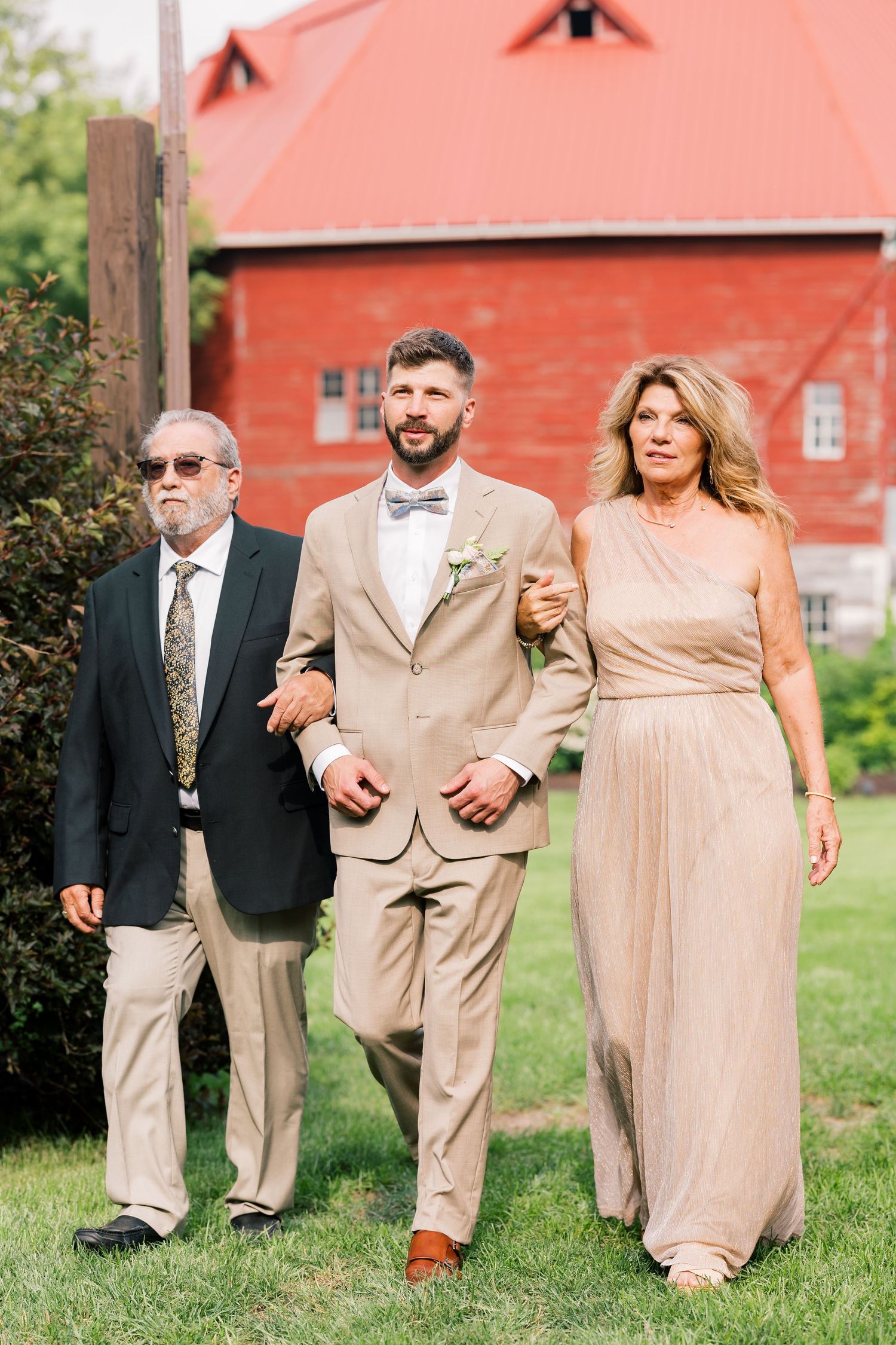 Wedding party in beige and neutral attire walking together on green lawn near red barn.