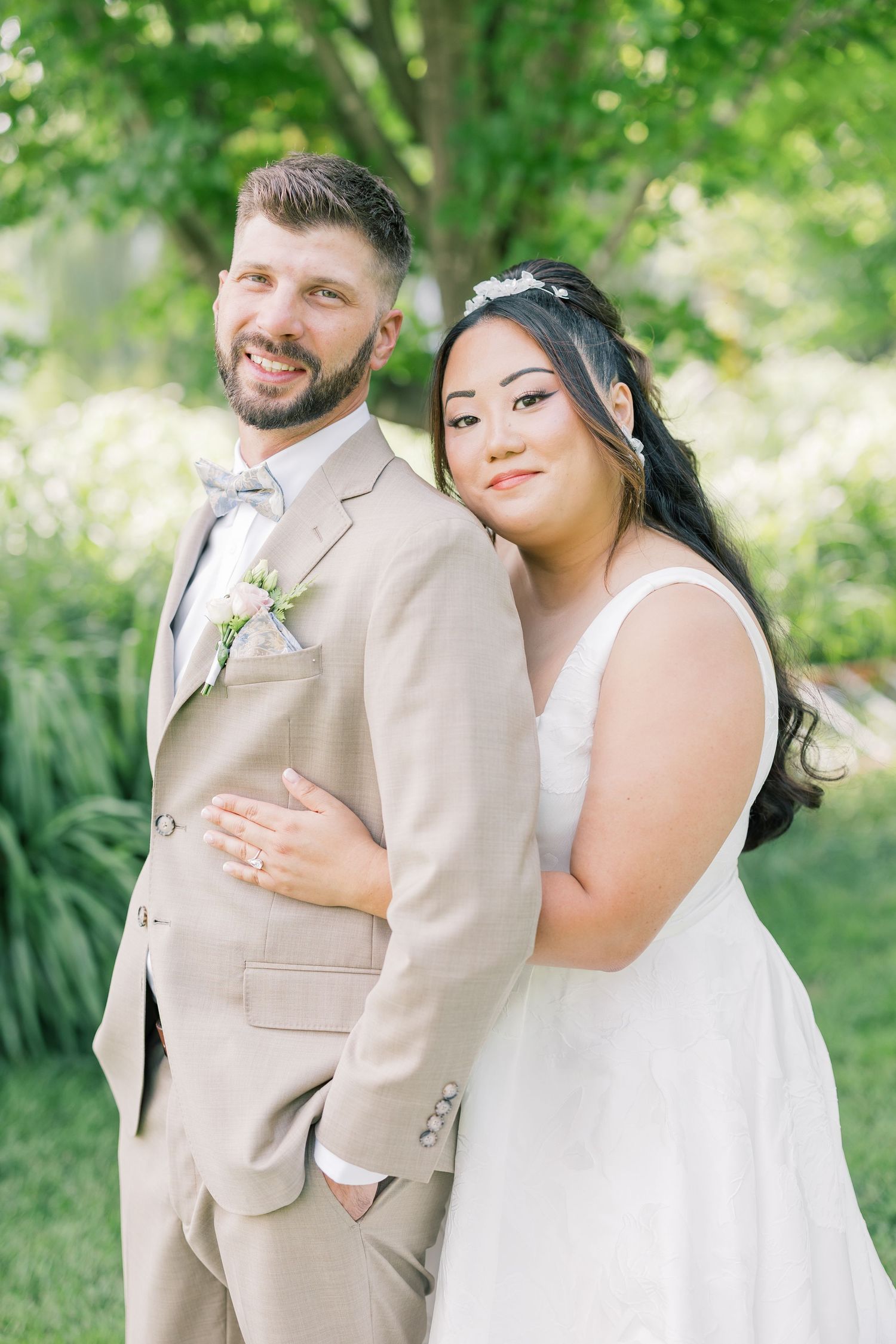 A romantic outdoor wedding portrait featuring a couple in tan suit and white dress against lush greenery.