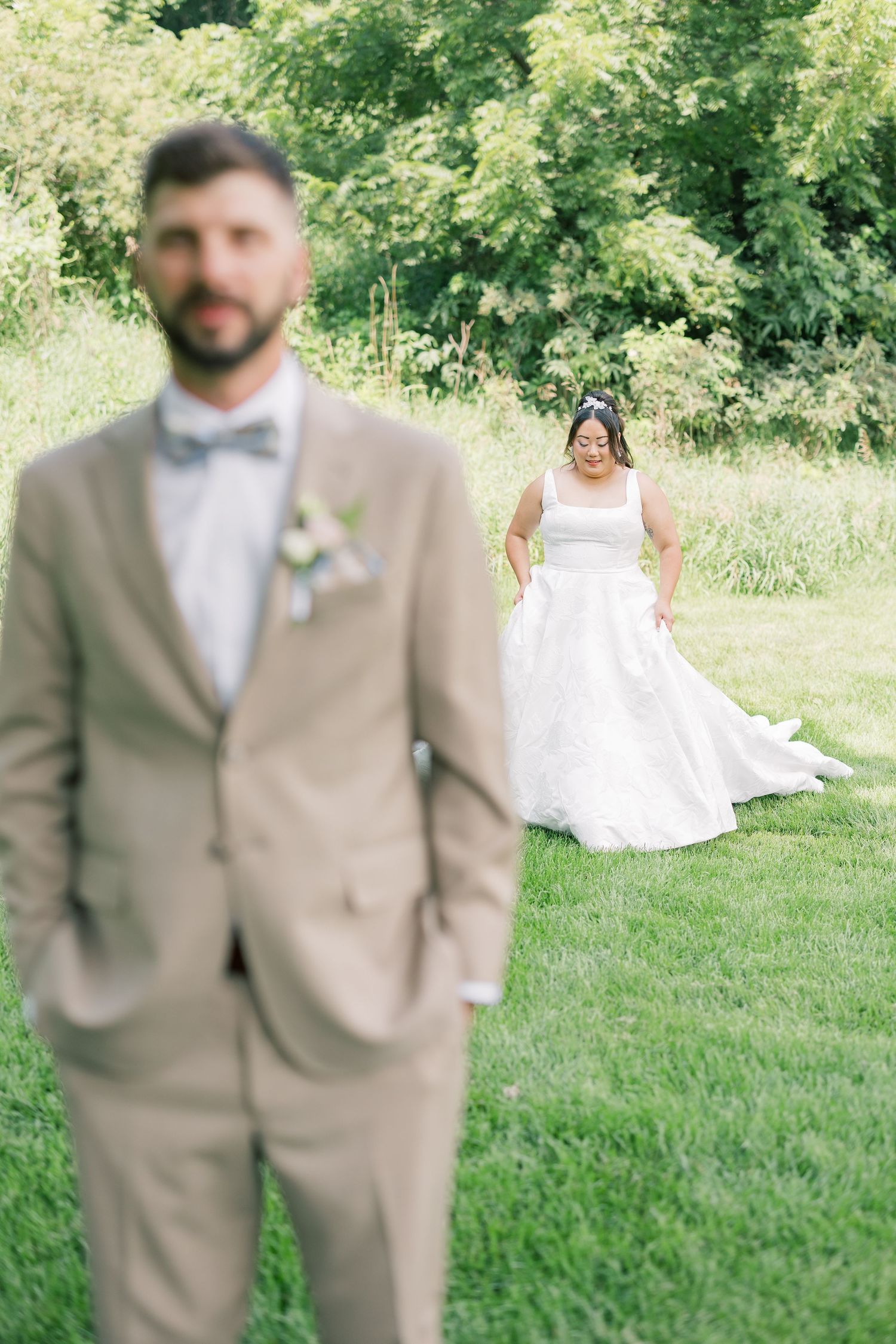A wedding first look moment with the bride in white standing behind the groom in a tan suit outdoors.