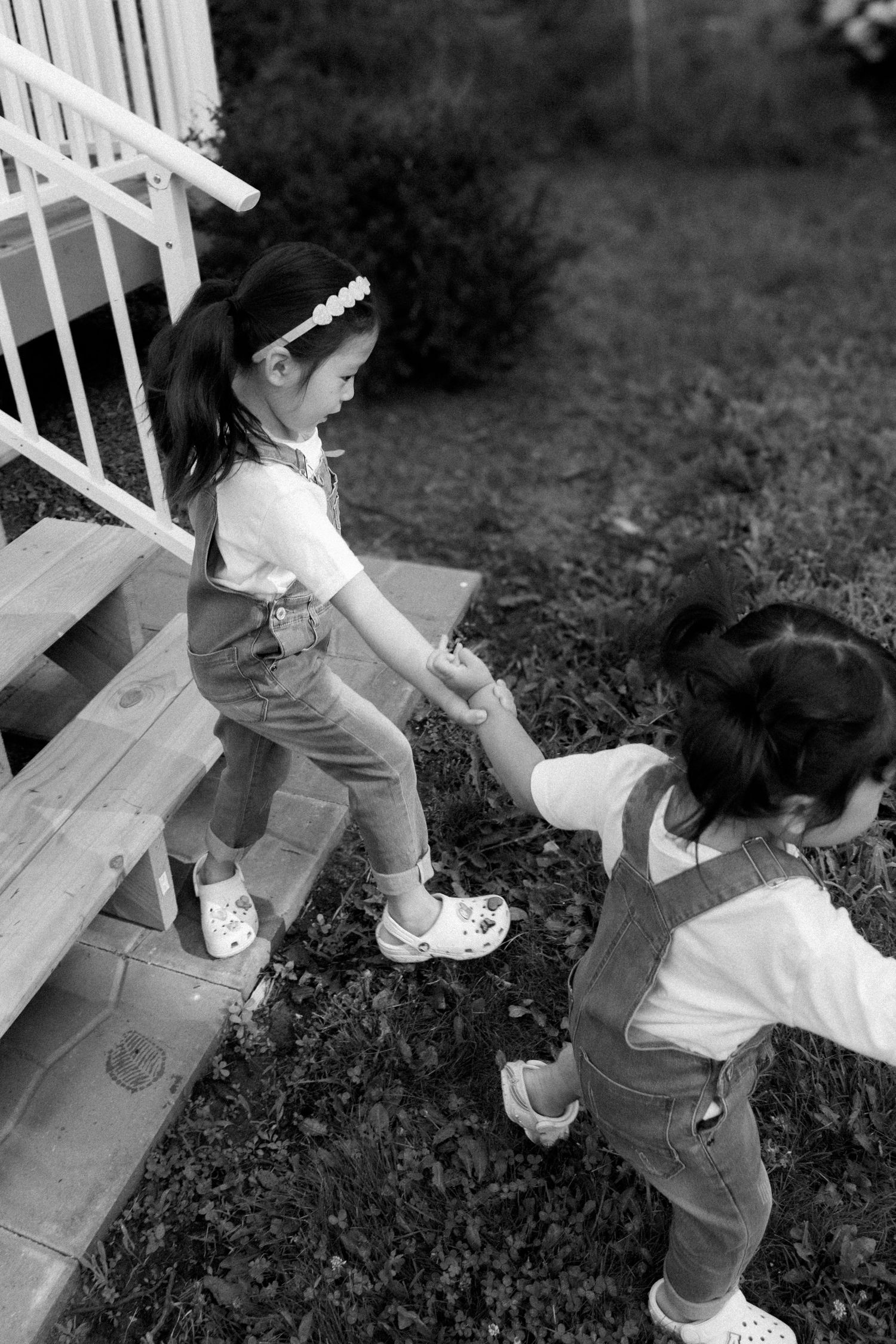 Two young girls in matching overalls hold hands on outdoor wooden steps in a black and white photo.