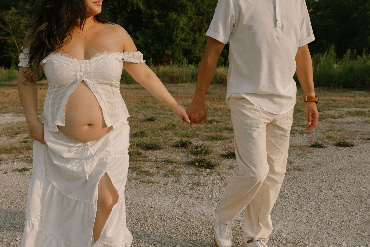 Pregnant woman in white off-shoulder dress holds hands with partner outdoors during maternity photoshoot.
