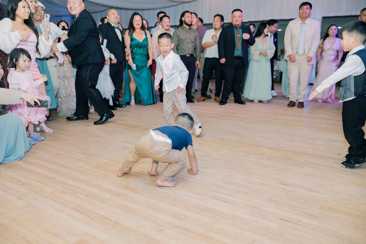 A young child breakdances on a wooden dance floor at a formal event while guests in formal attire watch from the sides.