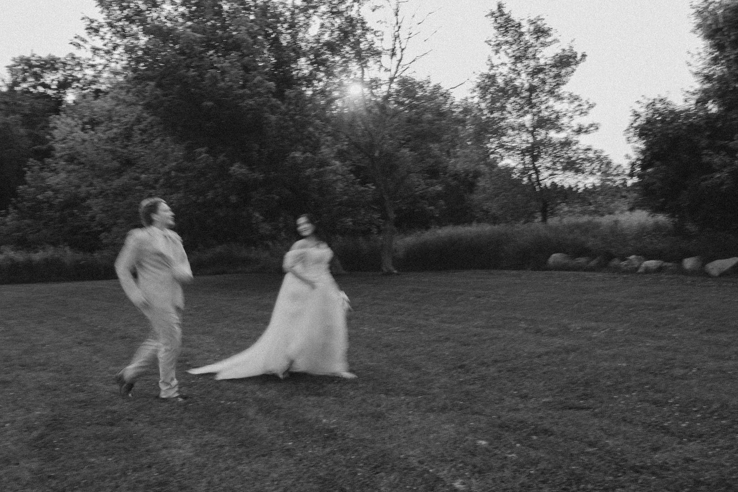 Black and white photo of a bride and groom running joyfully across a grassy field surrounded by trees.