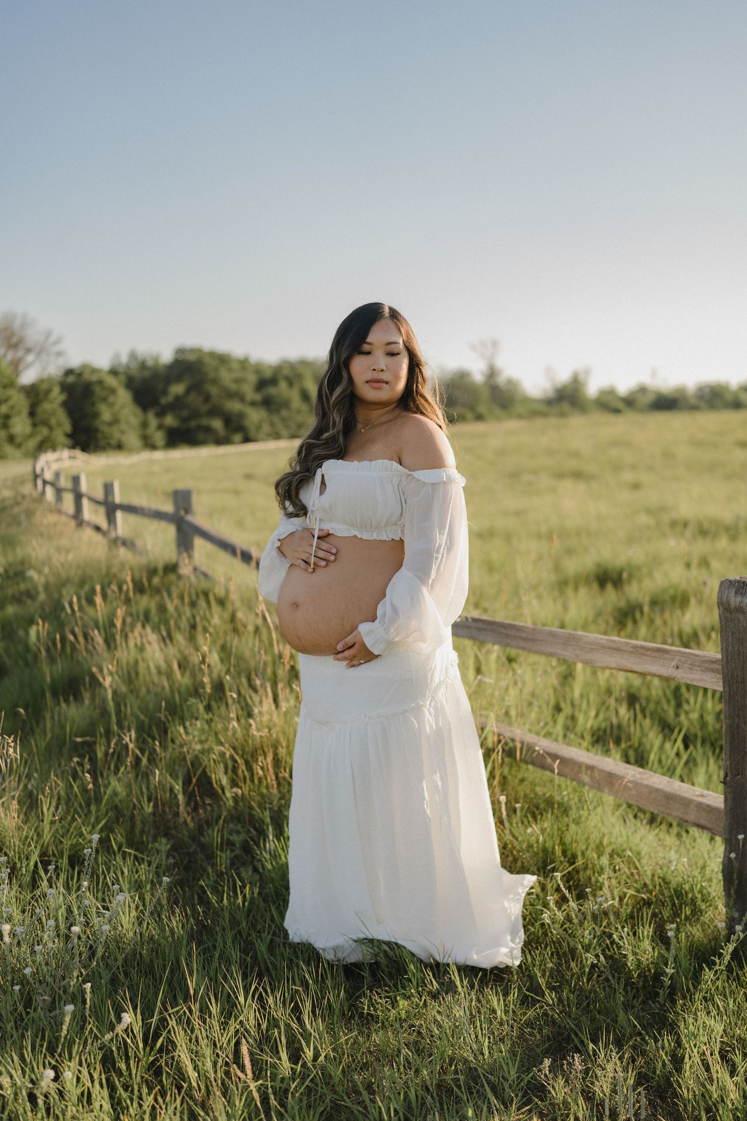 Pregnant woman in white off-shoulder gown cradling baby bump in sunny green pasture with wooden fence.