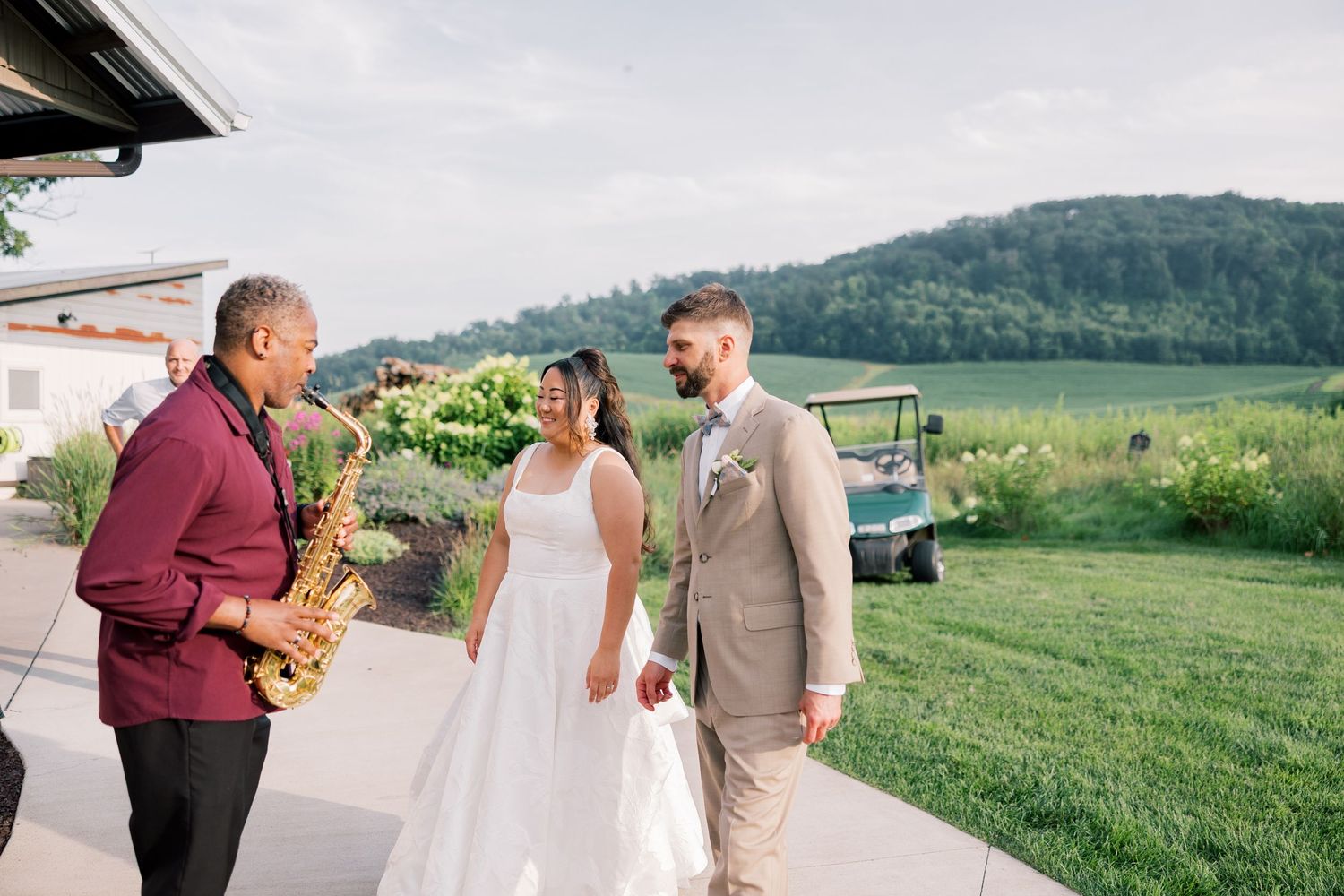 A wedding celebration takes place on a scenic hillside with green rolling mountains in the background.