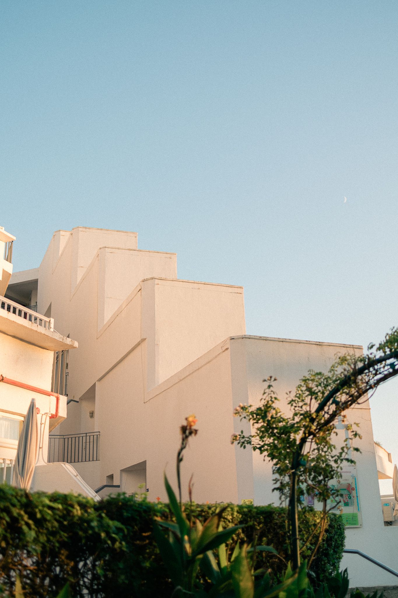 White Mediterranean-style building with geometric architecture against clear blue sky, surrounded by lush green plants.