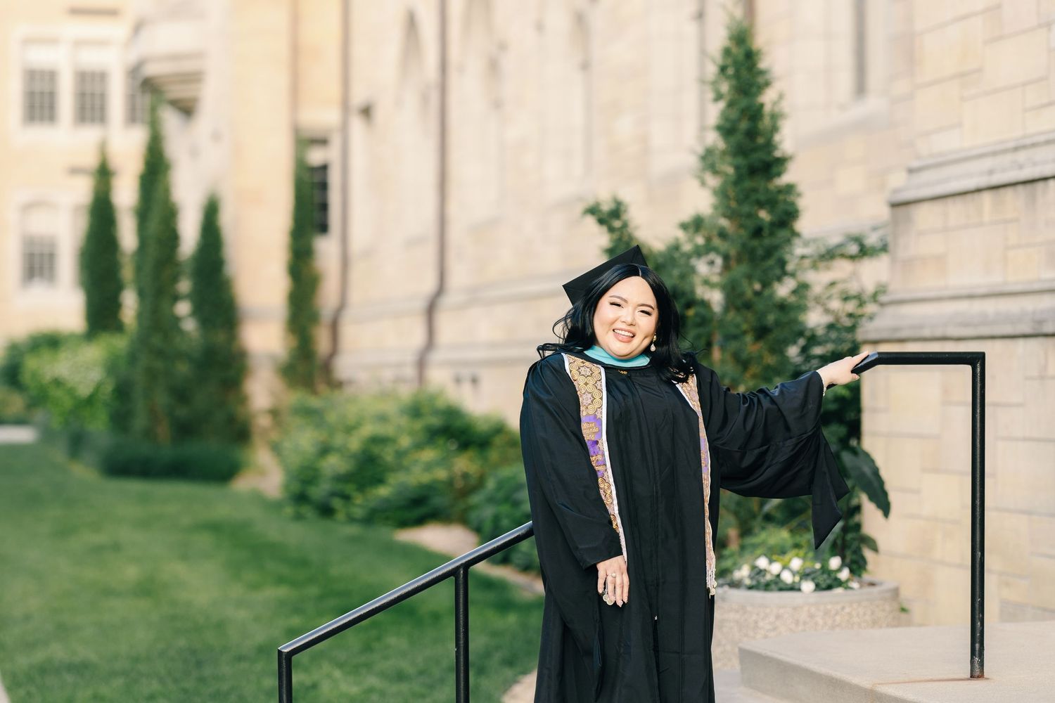 Graduate poses in traditional black commencement attire beside stone building with cypress trees.