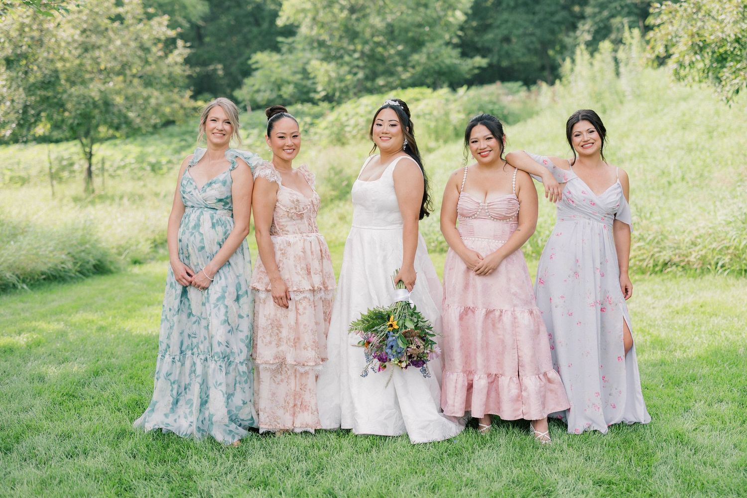 Wedding party in pastel dresses poses on a grassy hillside.