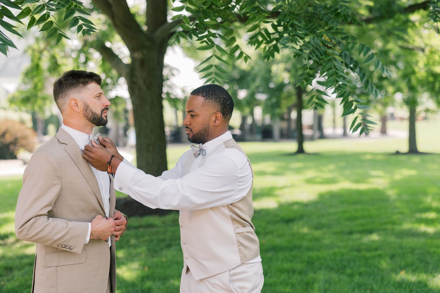 Two people in beige and white outfits share a playful moment on a grassy lawn under trees.