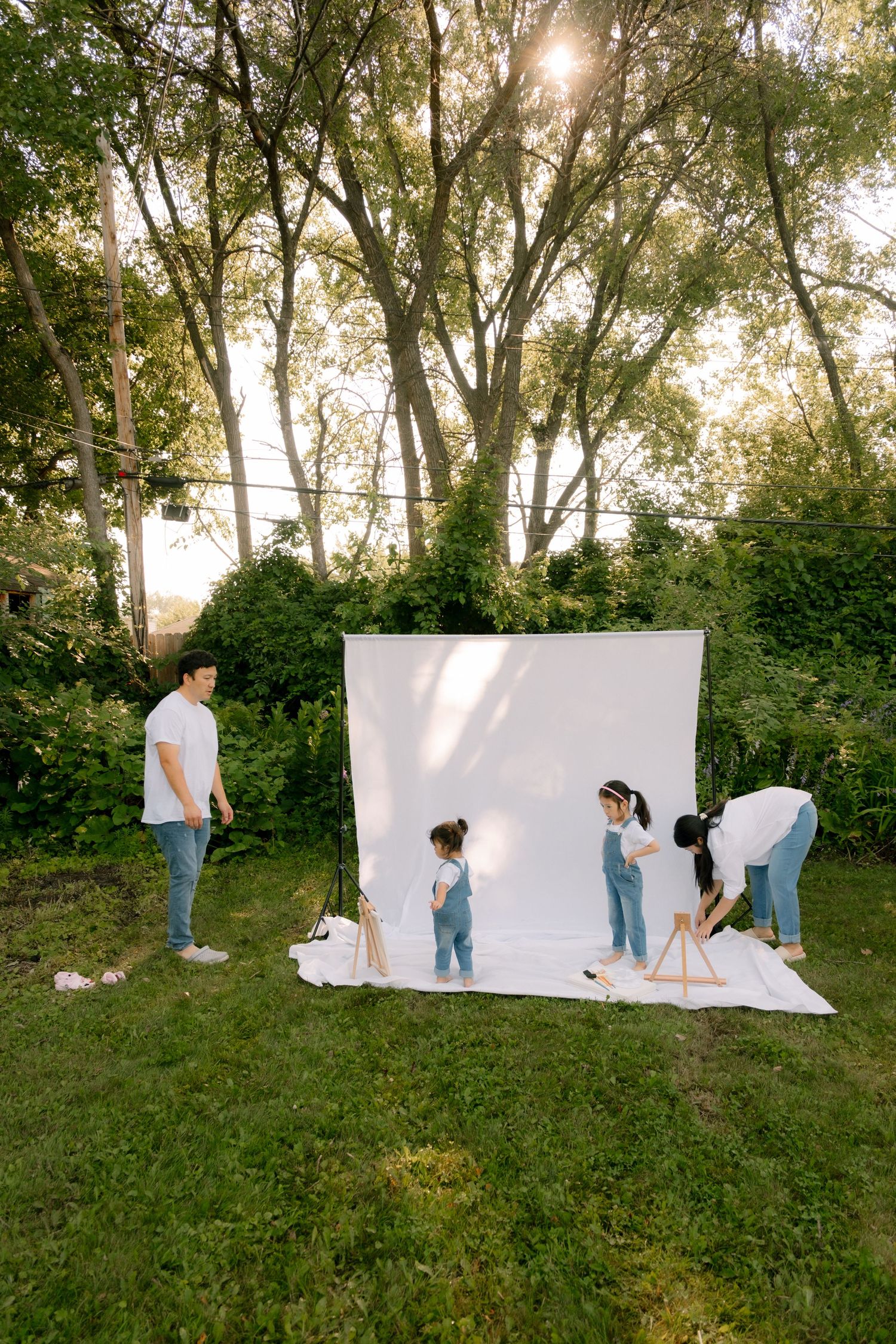 Family setting up a white backdrop for an outdoor photo shoot in a lush green backyard on a sunny day.