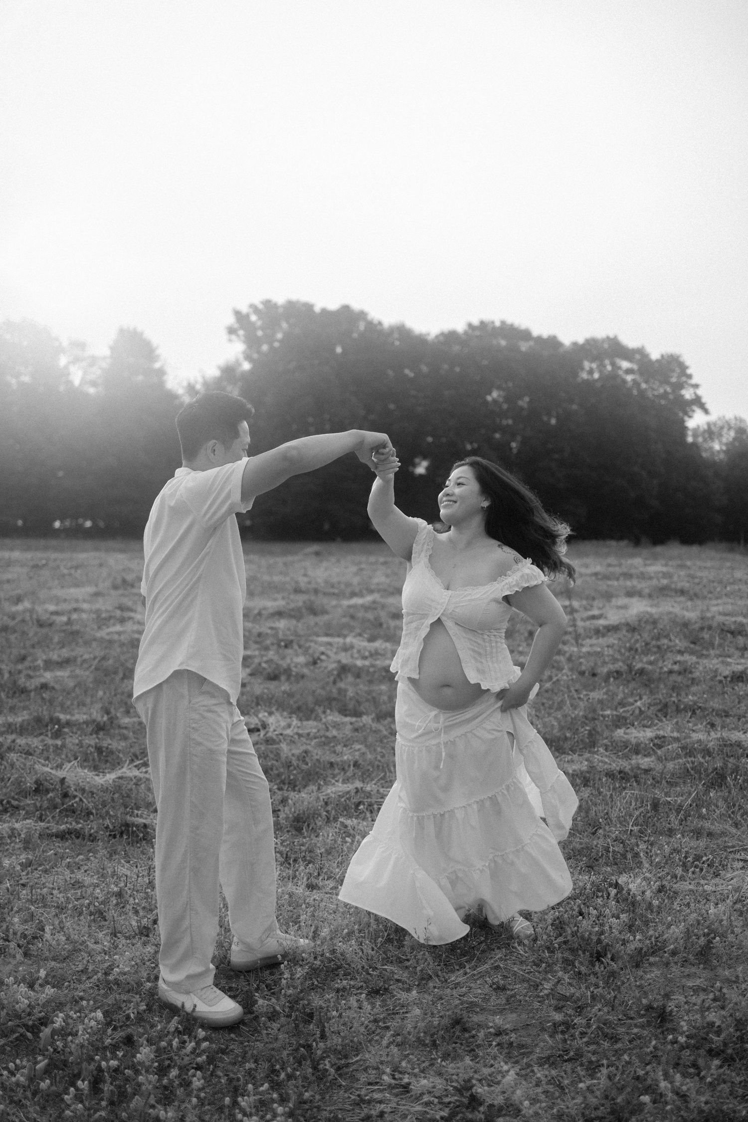 Black and white photo of a couple dancing in a field, pregnant woman in white dress being twirled by her partner.