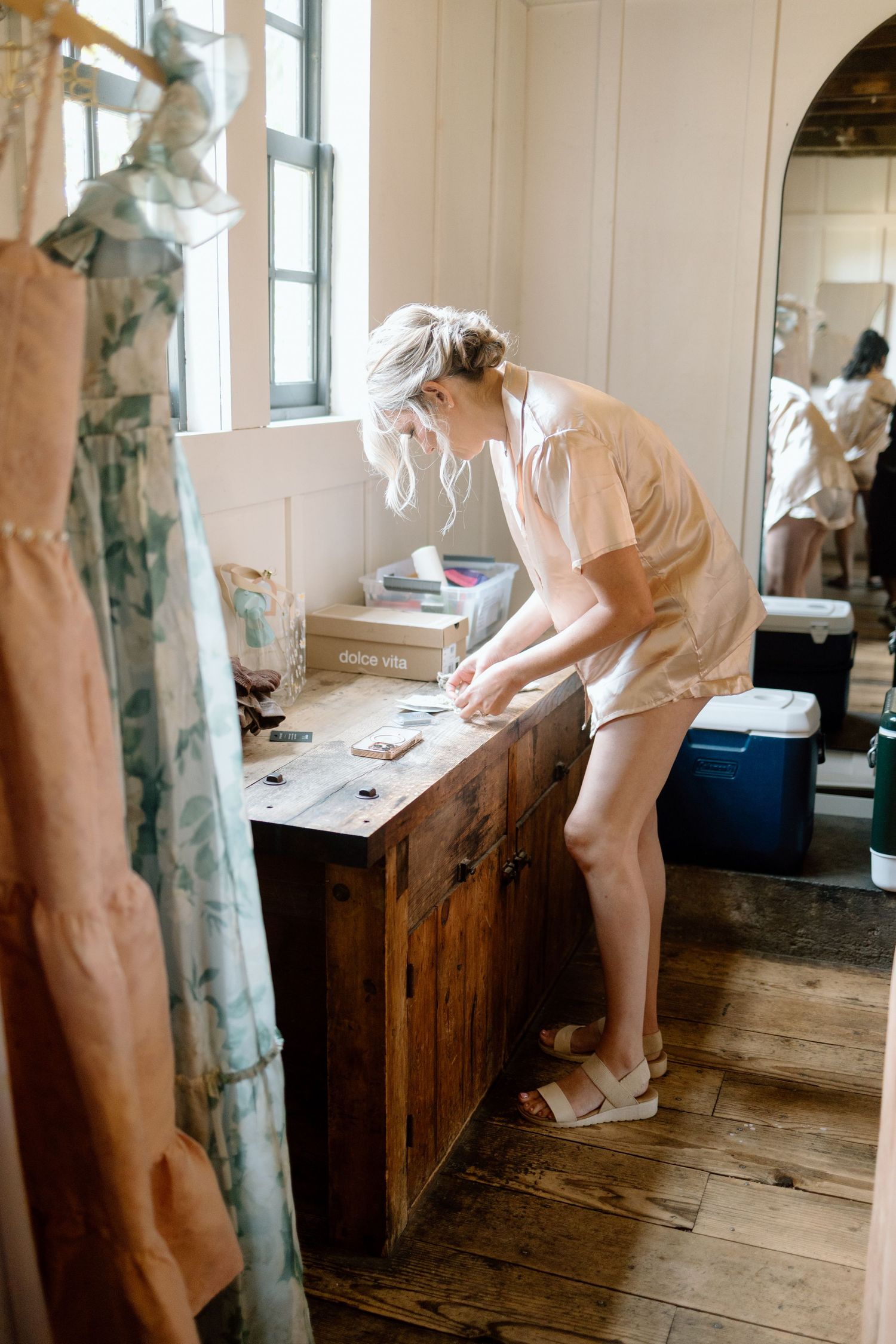 A person in a white dress bends over a wooden desk in a rustic room with windows.