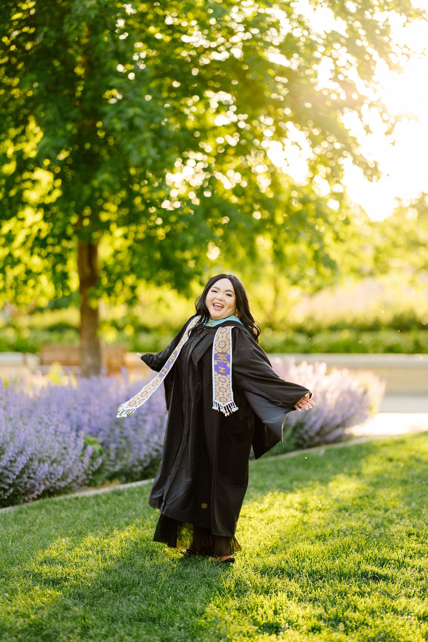 Graduate in black cap and gown poses joyfully in a garden with blooming lavender and sunlit trees.