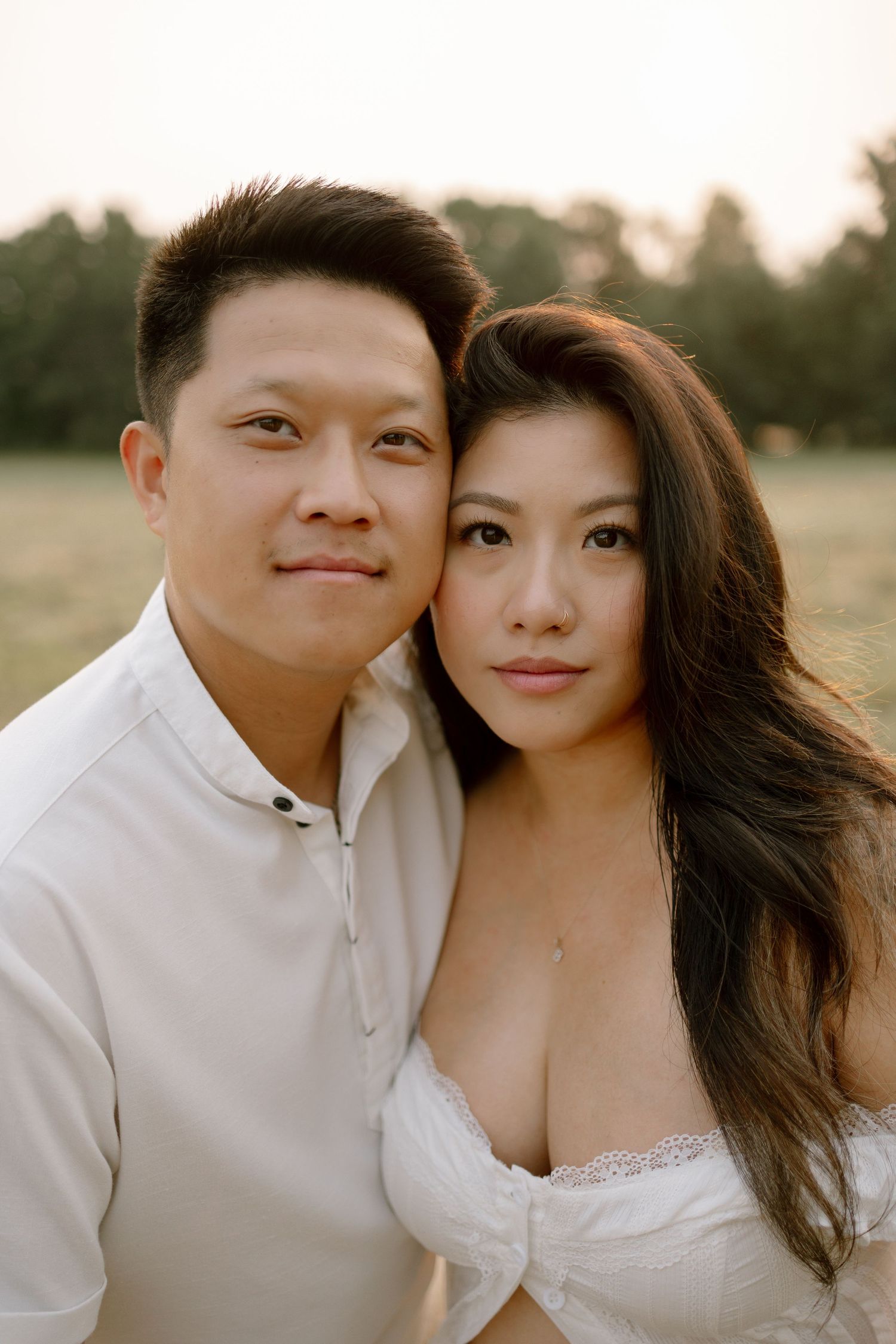 A couple in white outfits poses closely together in a golden field at sunset, smiling softly at the camera.