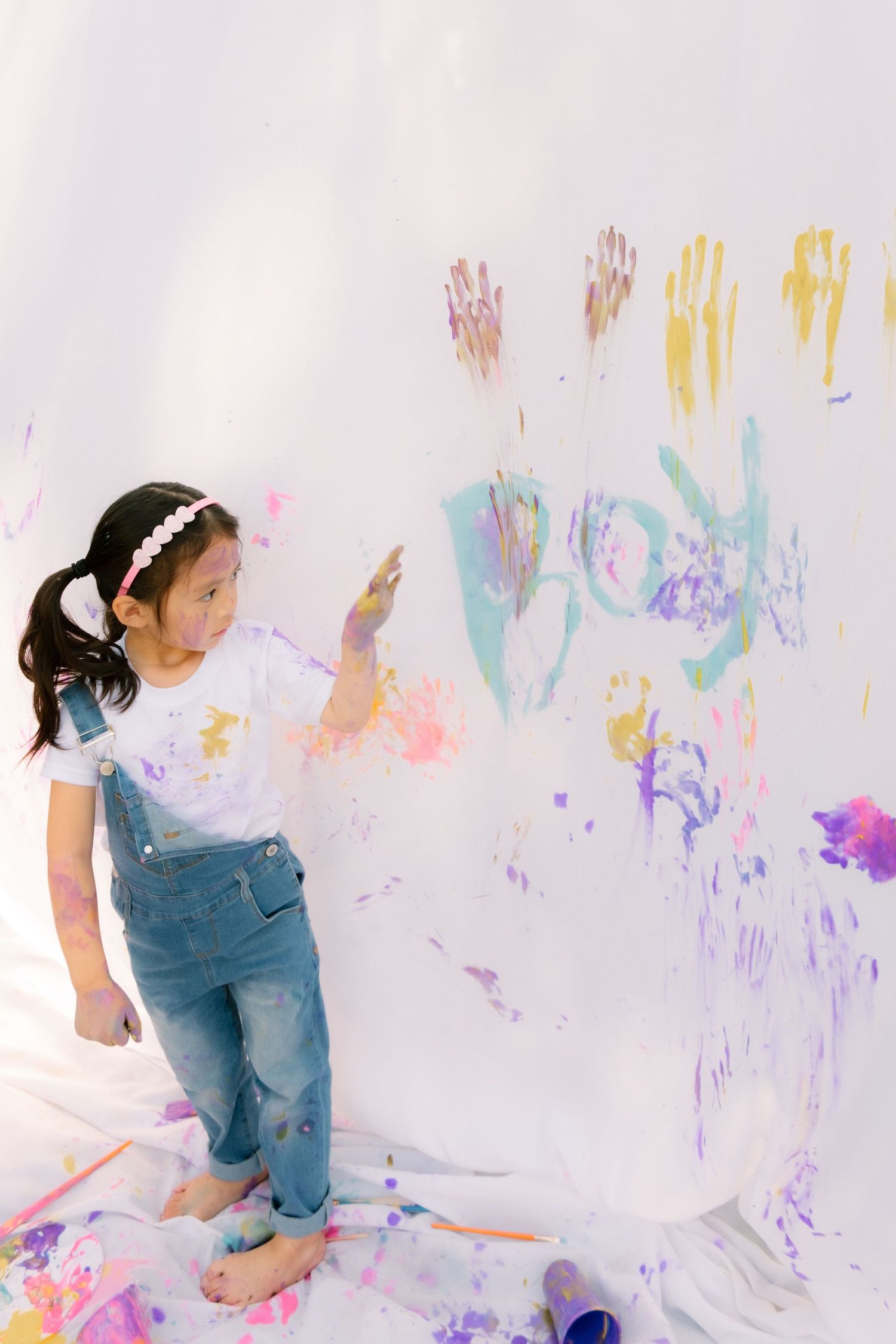 Young Asian girl in denim overalls creates colorful handprint art on large white backdrop during outdoor painting activity.