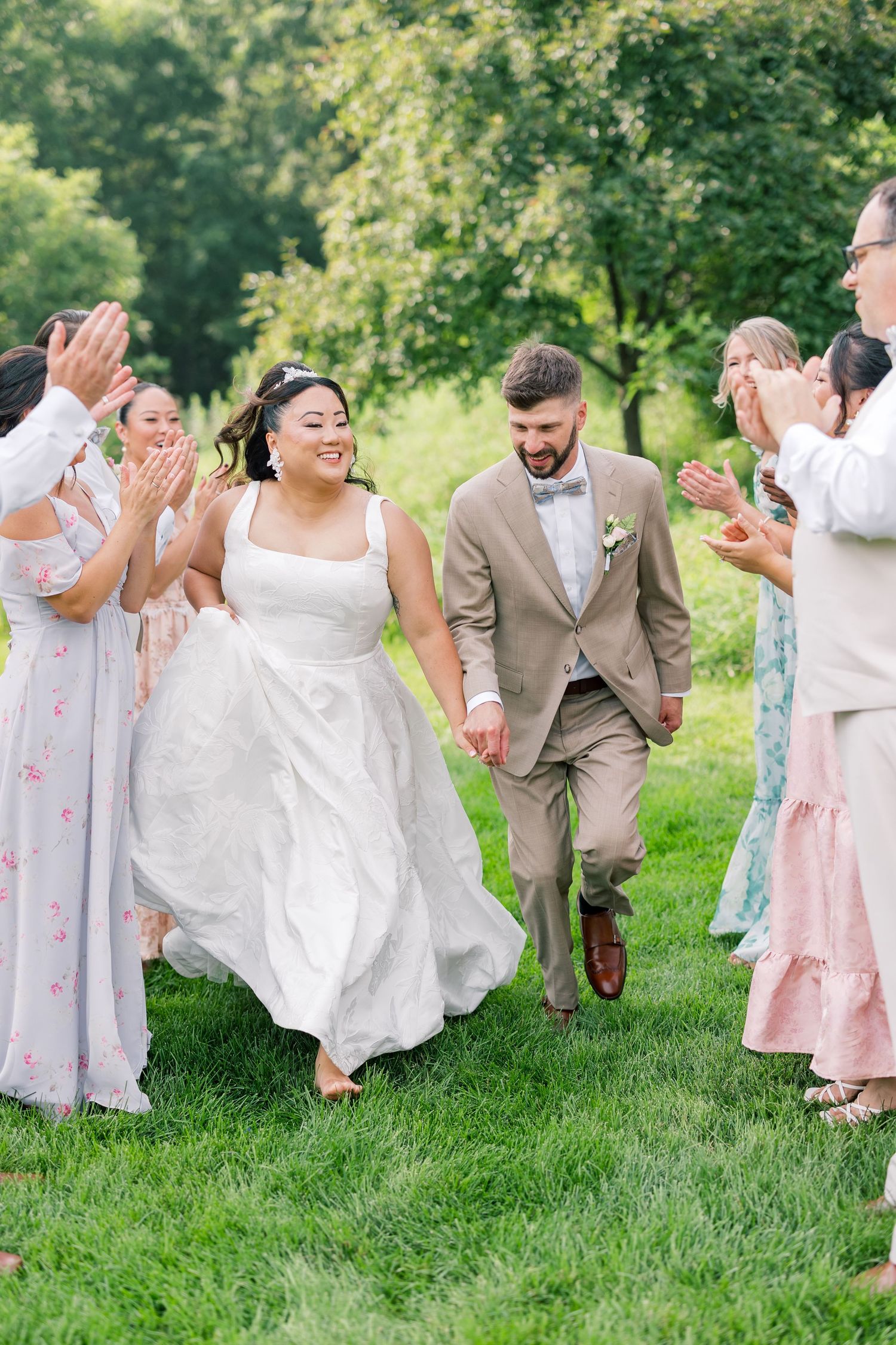 Wedding party in beige suits and floral dresses poses playfully on green grass.