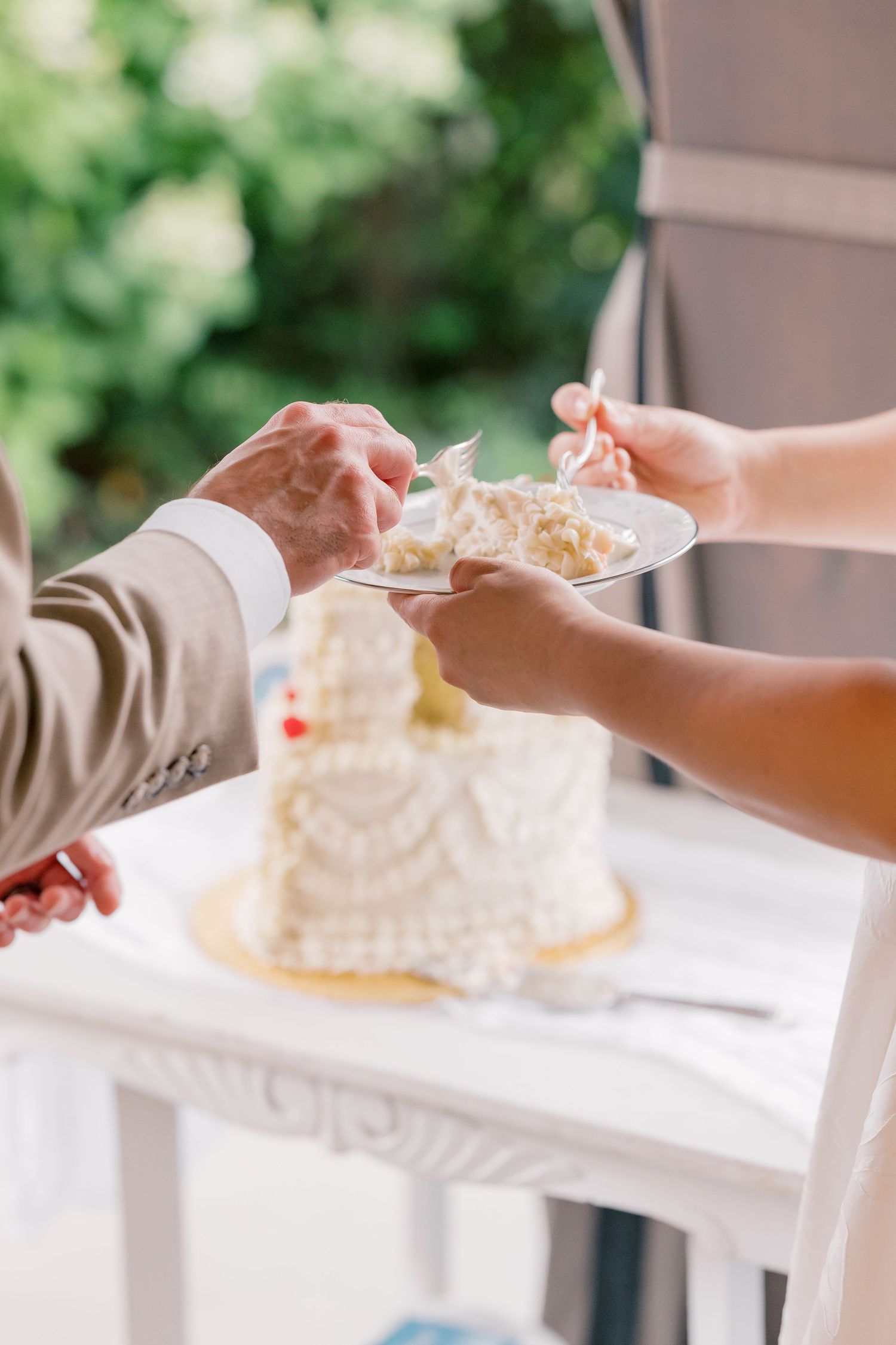 Hands serving slices of white wedding cake on elegant plates at an outdoor reception.
