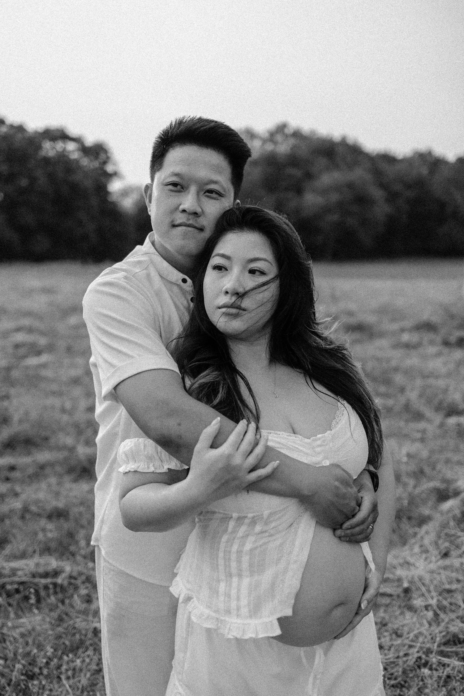 Black and white maternity photo of a couple embracing in a field, woman in white dress cradling baby bump.
