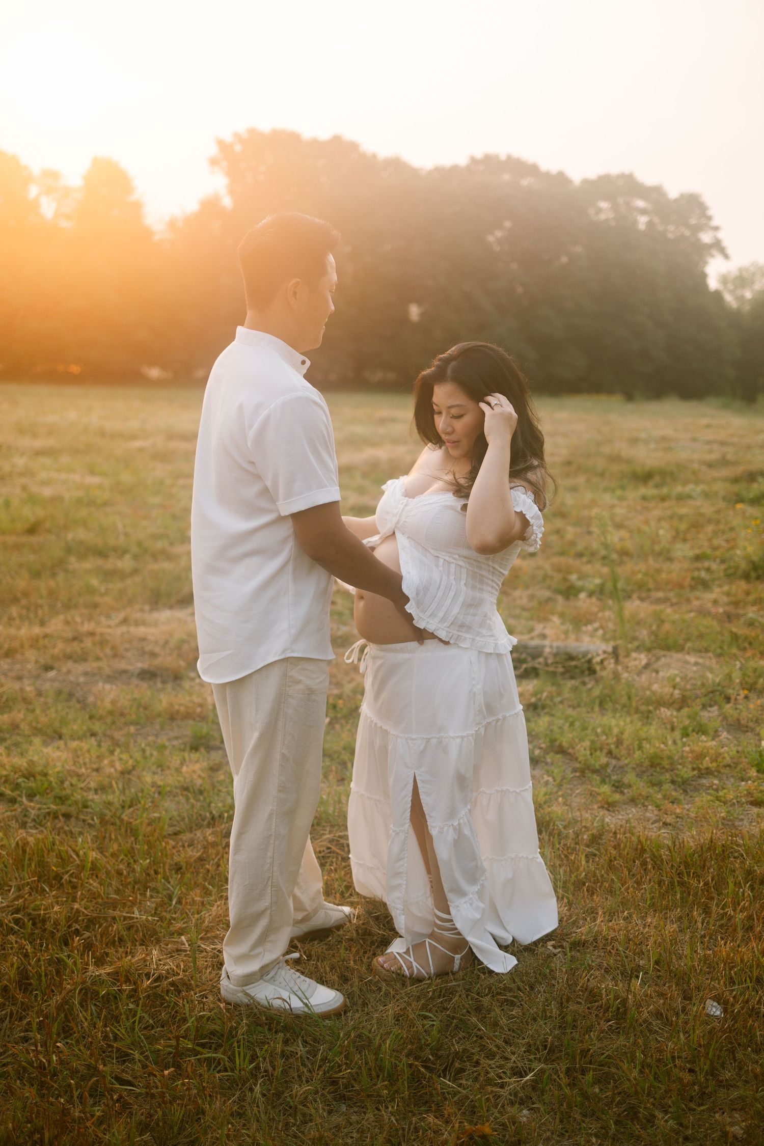 Couple in white outfits holding hands in a golden sunset field, woman in white dress touching her hair.