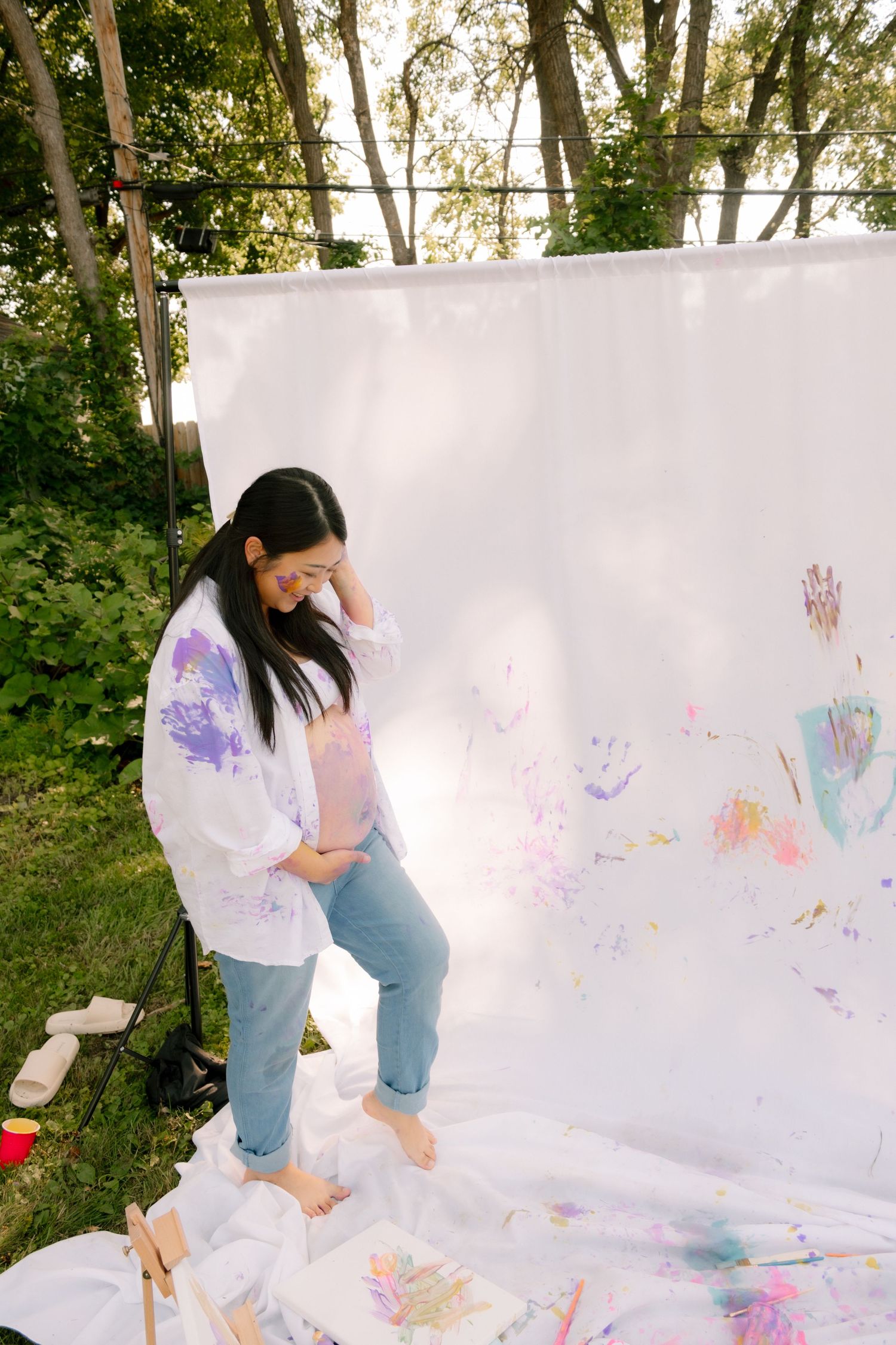 Pregnant woman in paint-splattered white shirt standing barefoot by colorful painted white backdrop.
