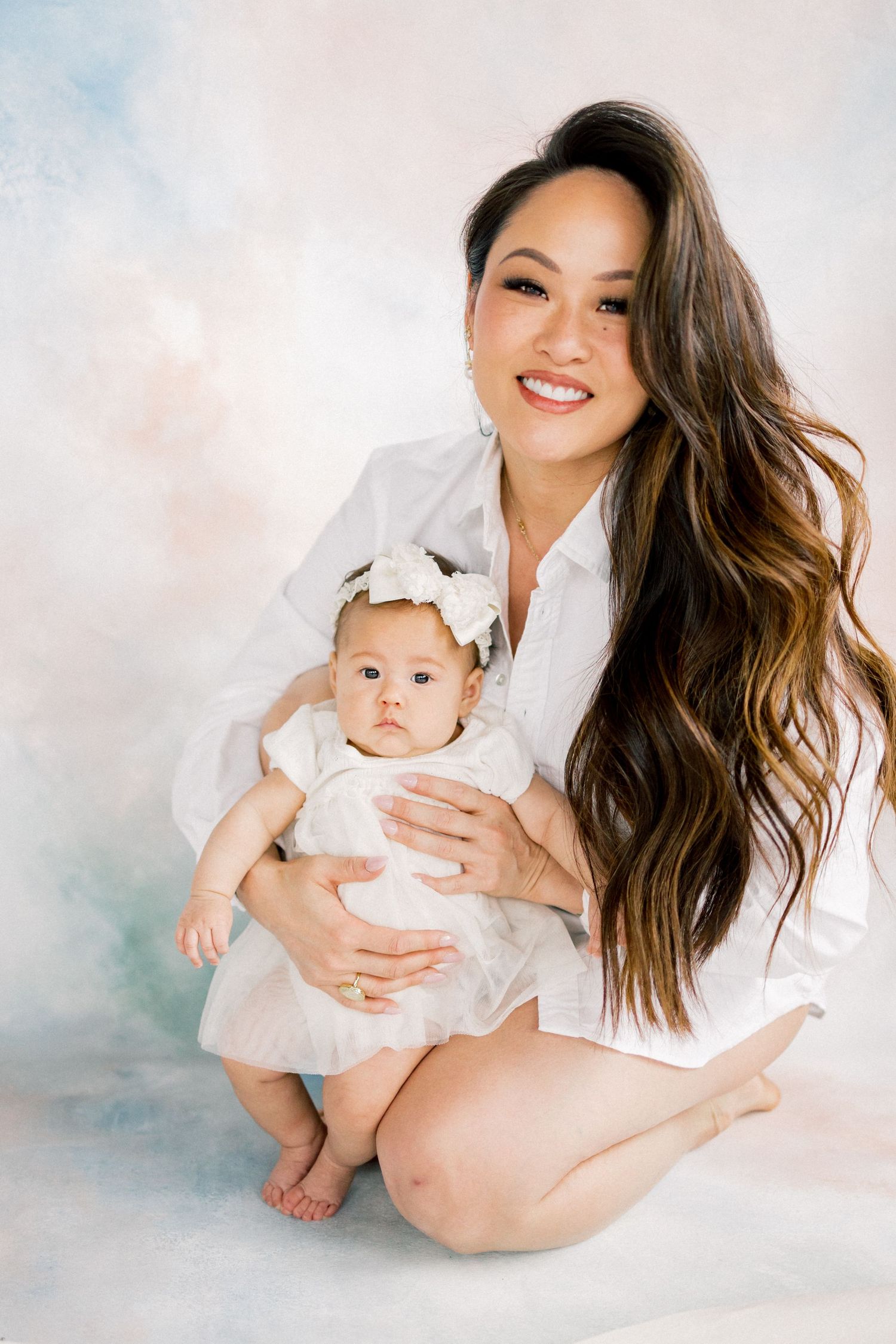Smiling woman with long hair holds a baby in white dress and bow headband against a soft pastel backdrop.