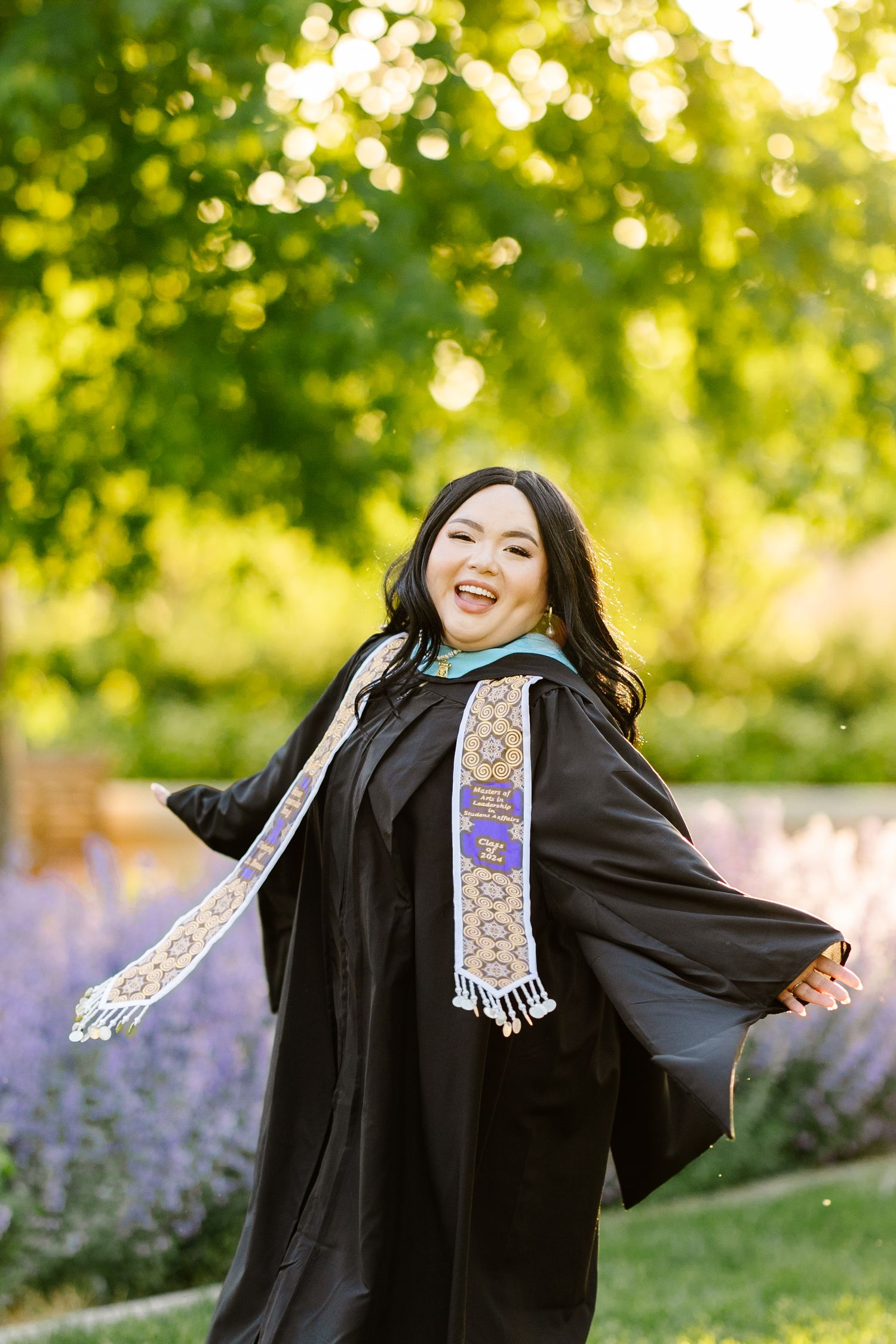 Graduate in black academic regalia poses joyfully against sunlit trees and purple flowers.