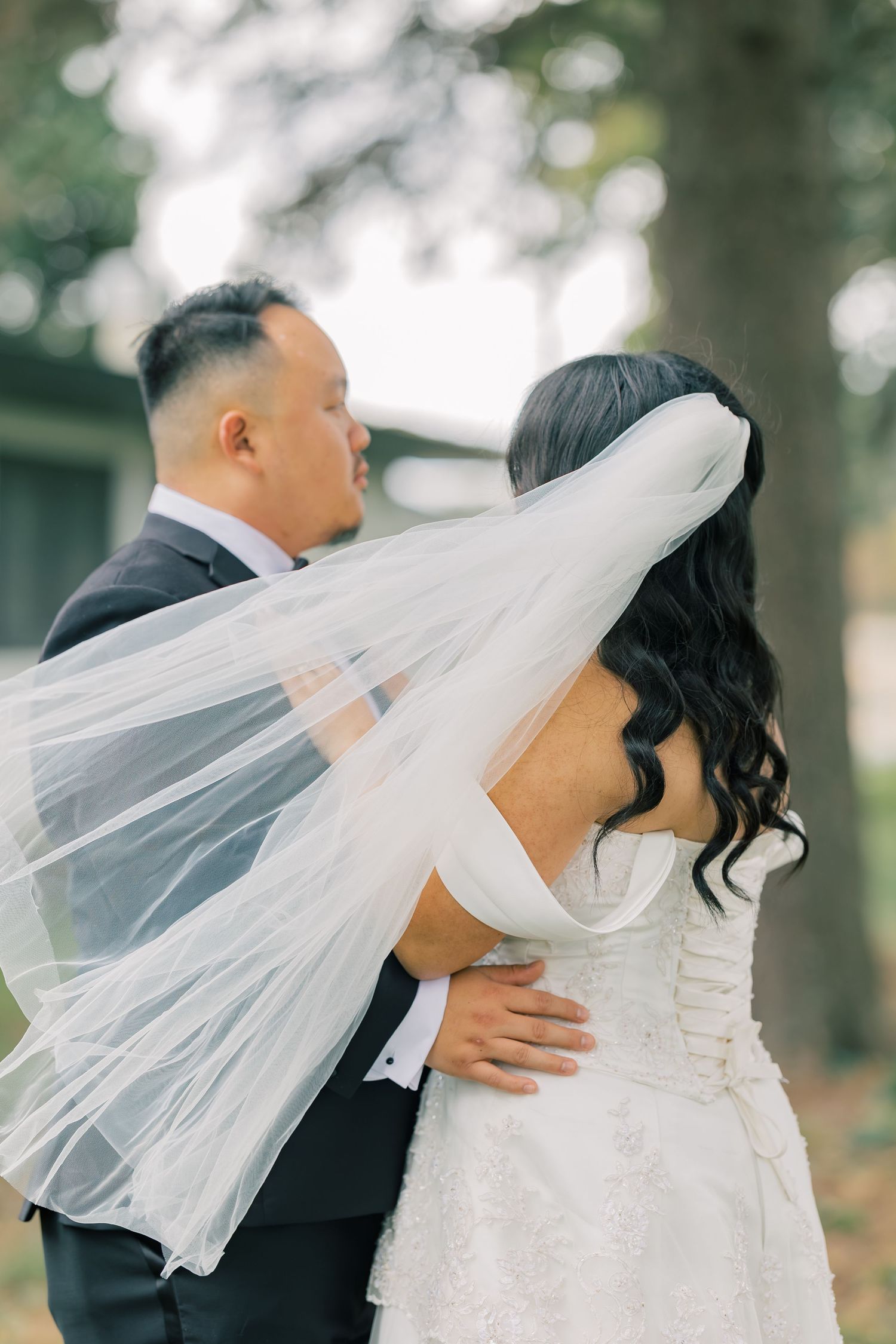 Romantic wedding couple embracing outdoors with flowing white veil in natural setting.