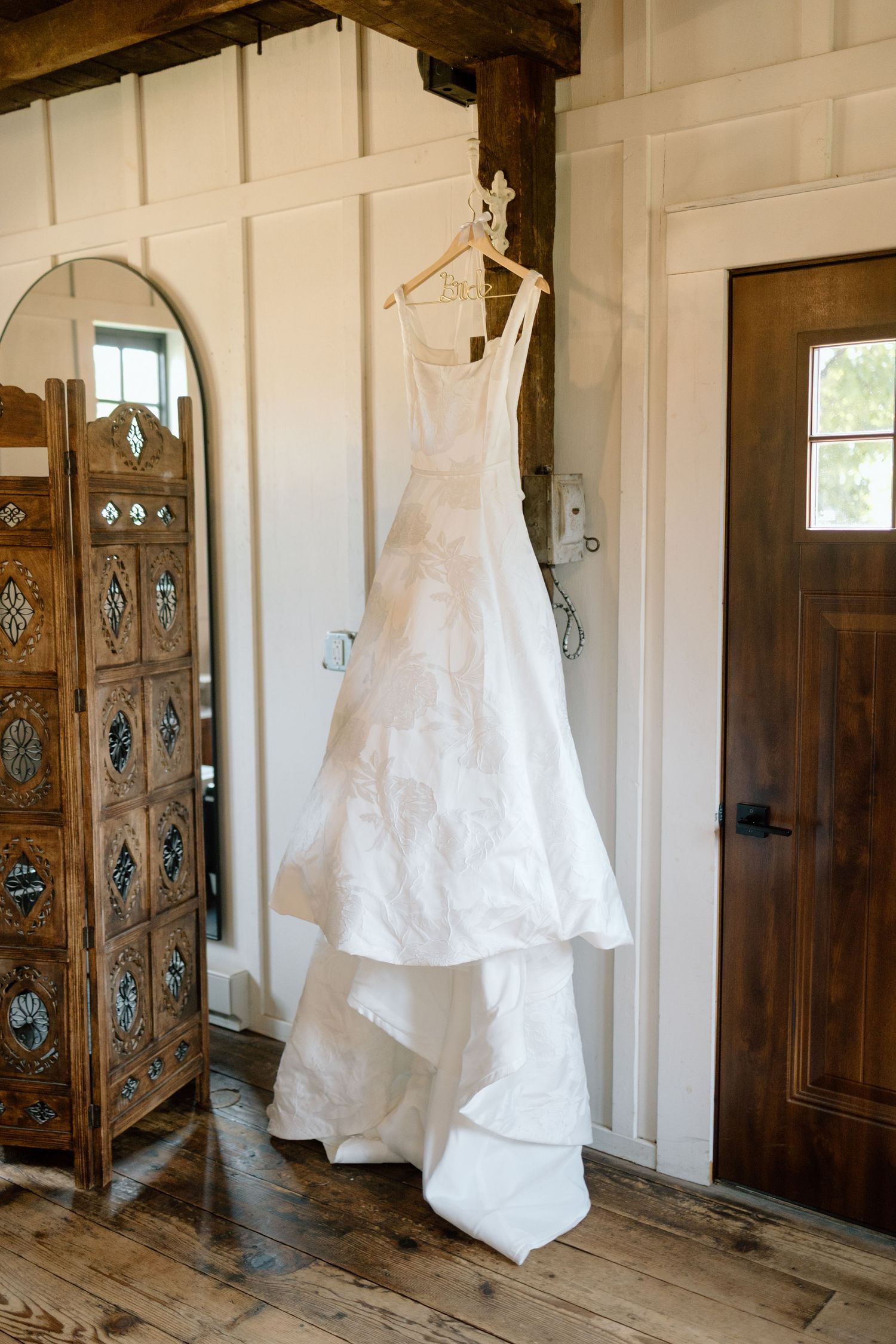 White wedding dress hanging in rustic wooden room with ornate carved doors.
