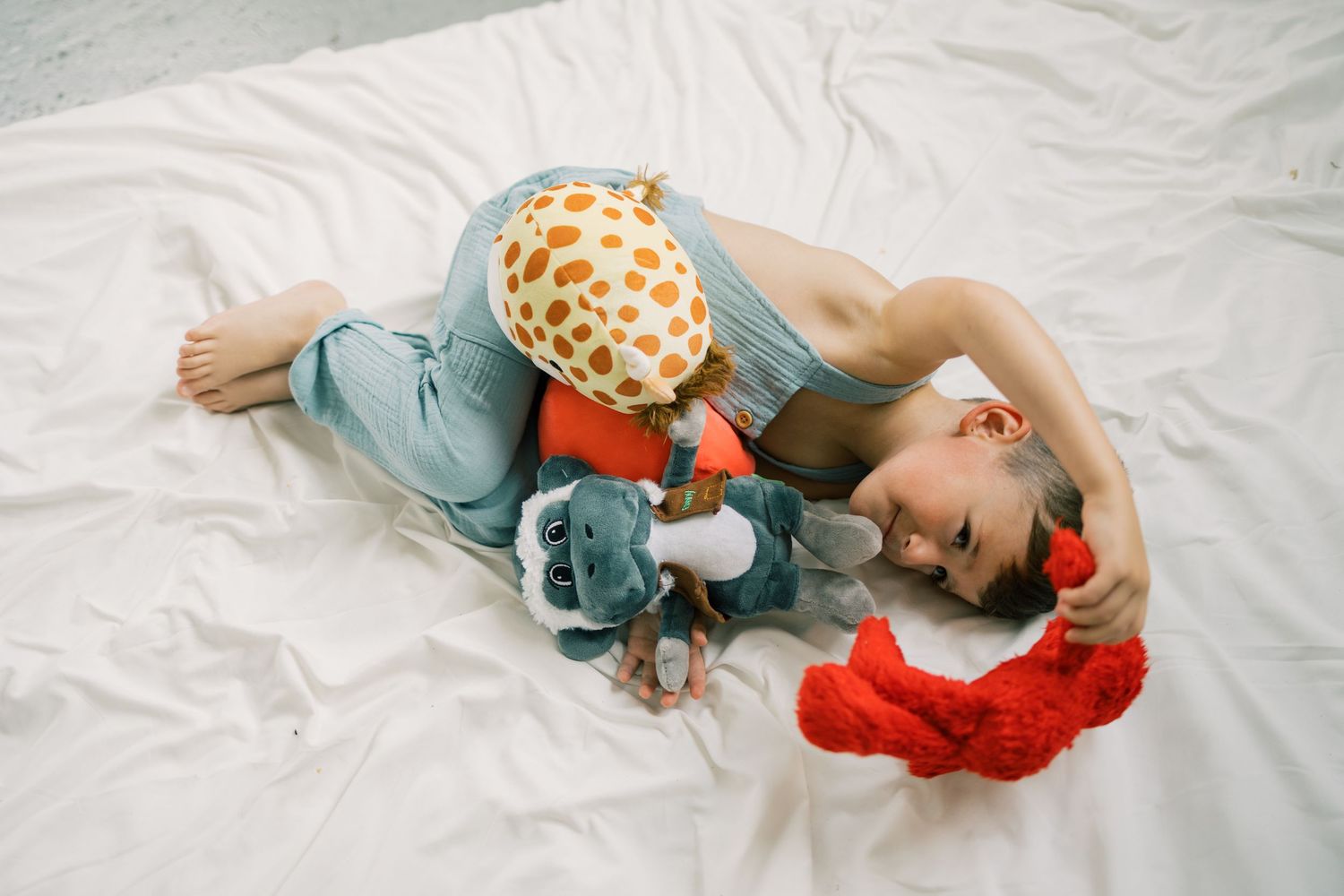 Child lying on white bedding smiling while hugging colorful stuffed animals including a giraffe and red toy.