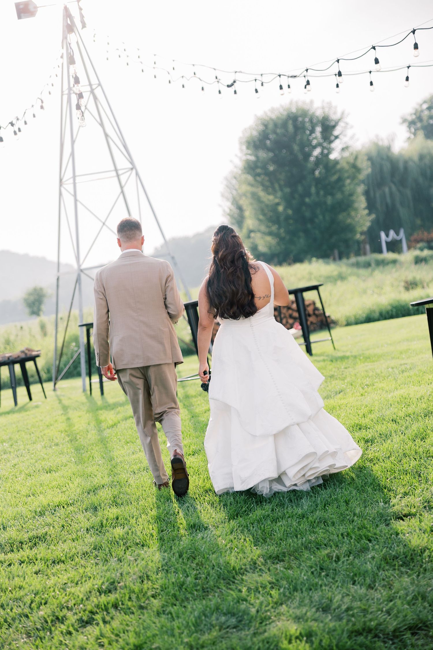 String lights hang above a grassy outdoor wedding venue as couple walks together.