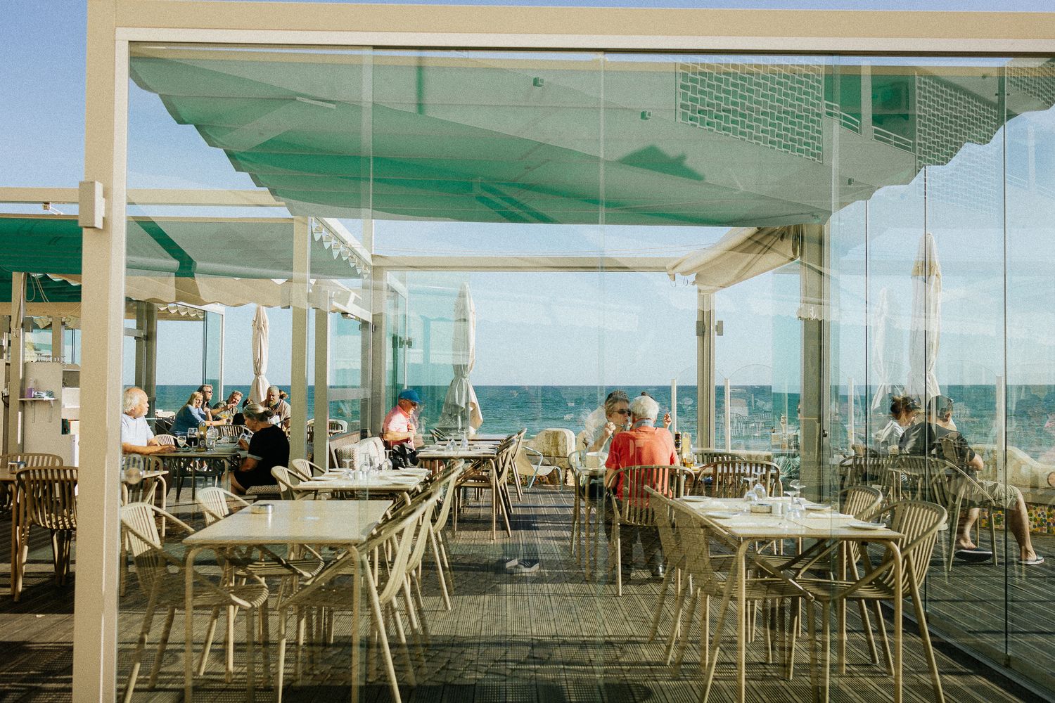 Beachfront restaurant with white tables and chairs under a turquoise awning overlooking the ocean on a sunny day.
