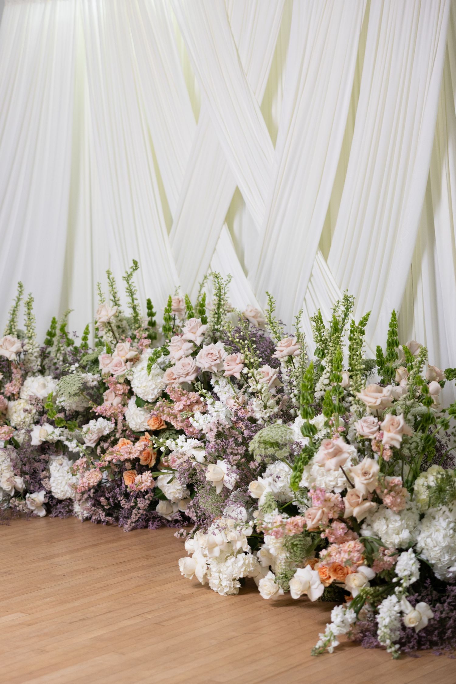 Elegant floral arrangement of pink and white flowers with greenery along a white draped backdrop on wooden flooring.