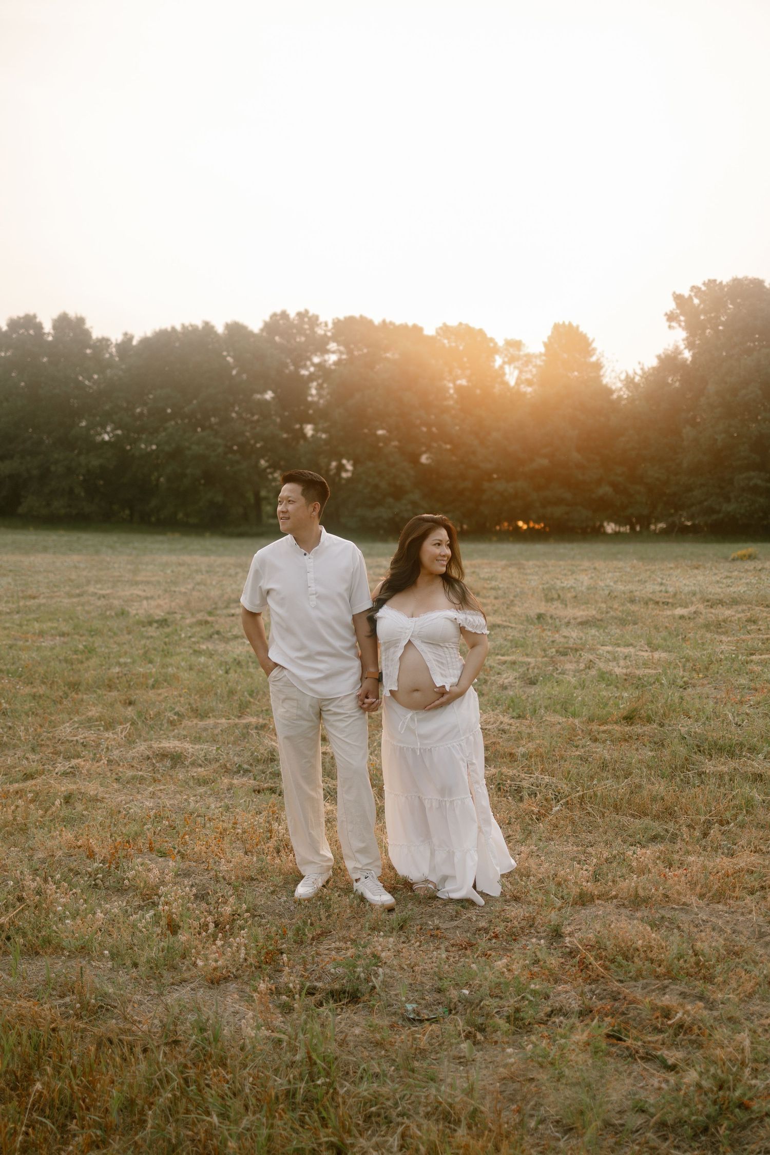 Pregnant couple in white outfits holding hands in a golden sunset field, maternity photography session outdoors.