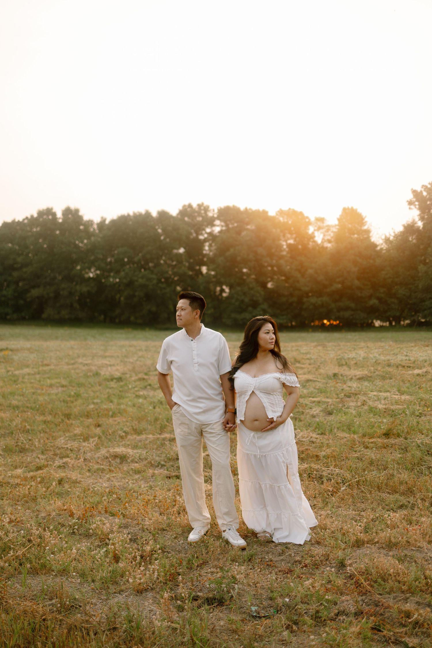 Expecting couple in matching white outfits holding hands in a sunlit field during golden hour maternity photoshoot.