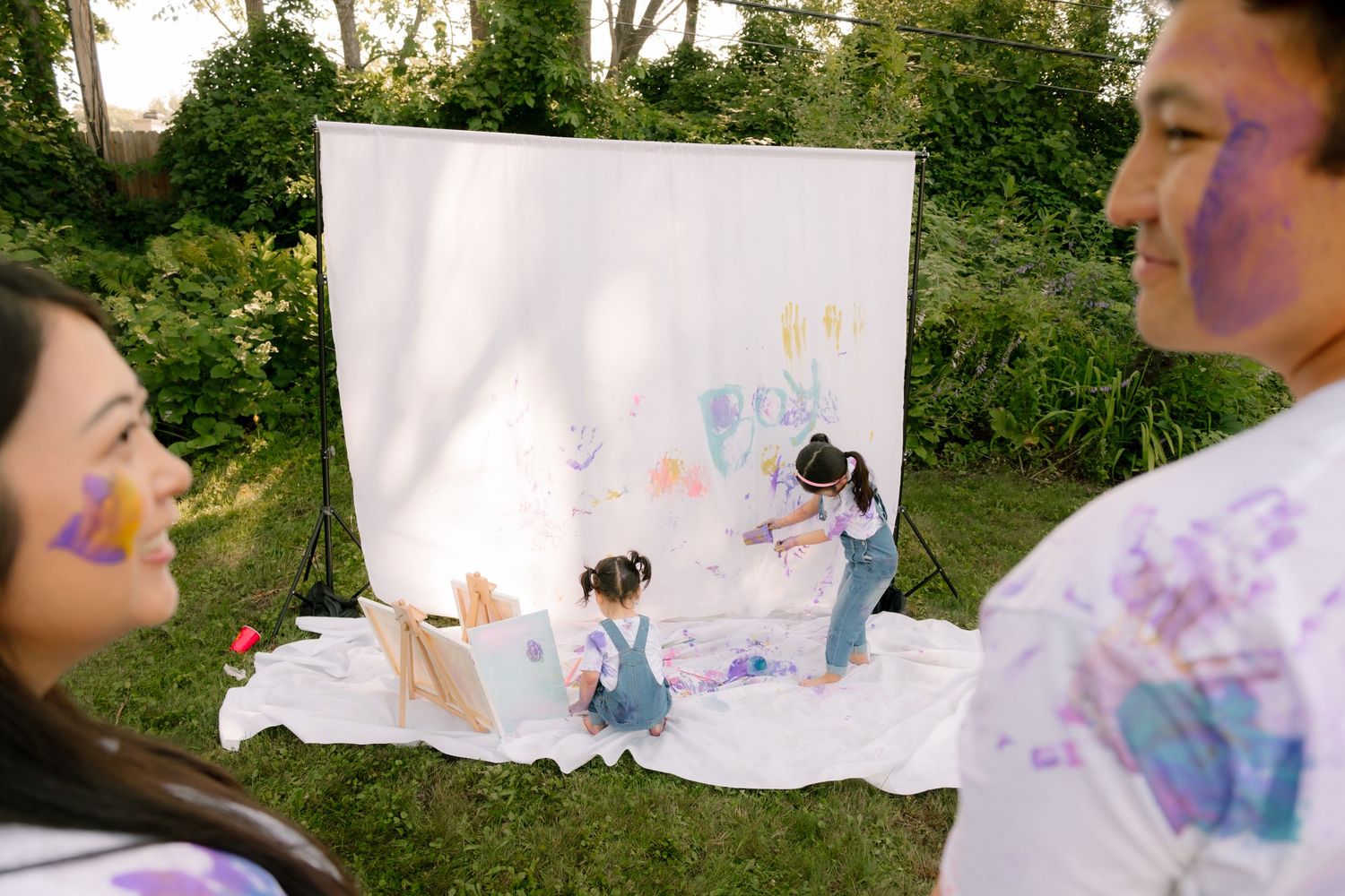 Family enjoying outdoor painting activity on white canvas backdrop in garden, with paint-covered faces and clothes.