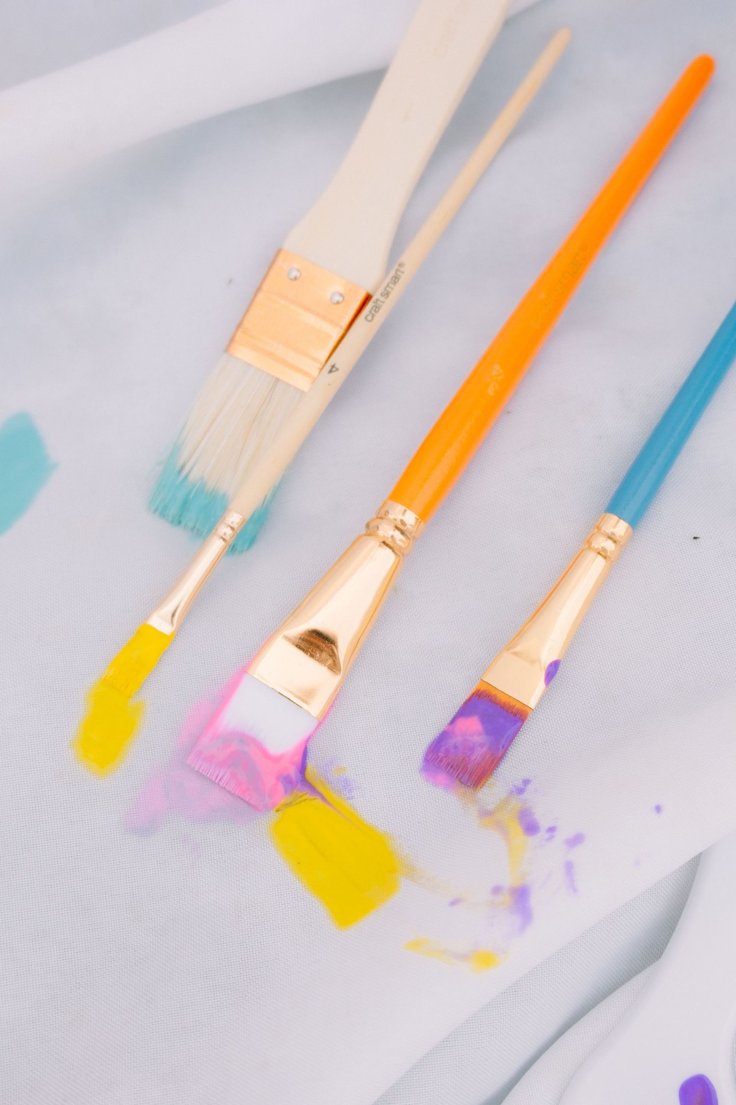 Three colorful paint brushes with yellow, purple, and blue paint strokes on white fabric.